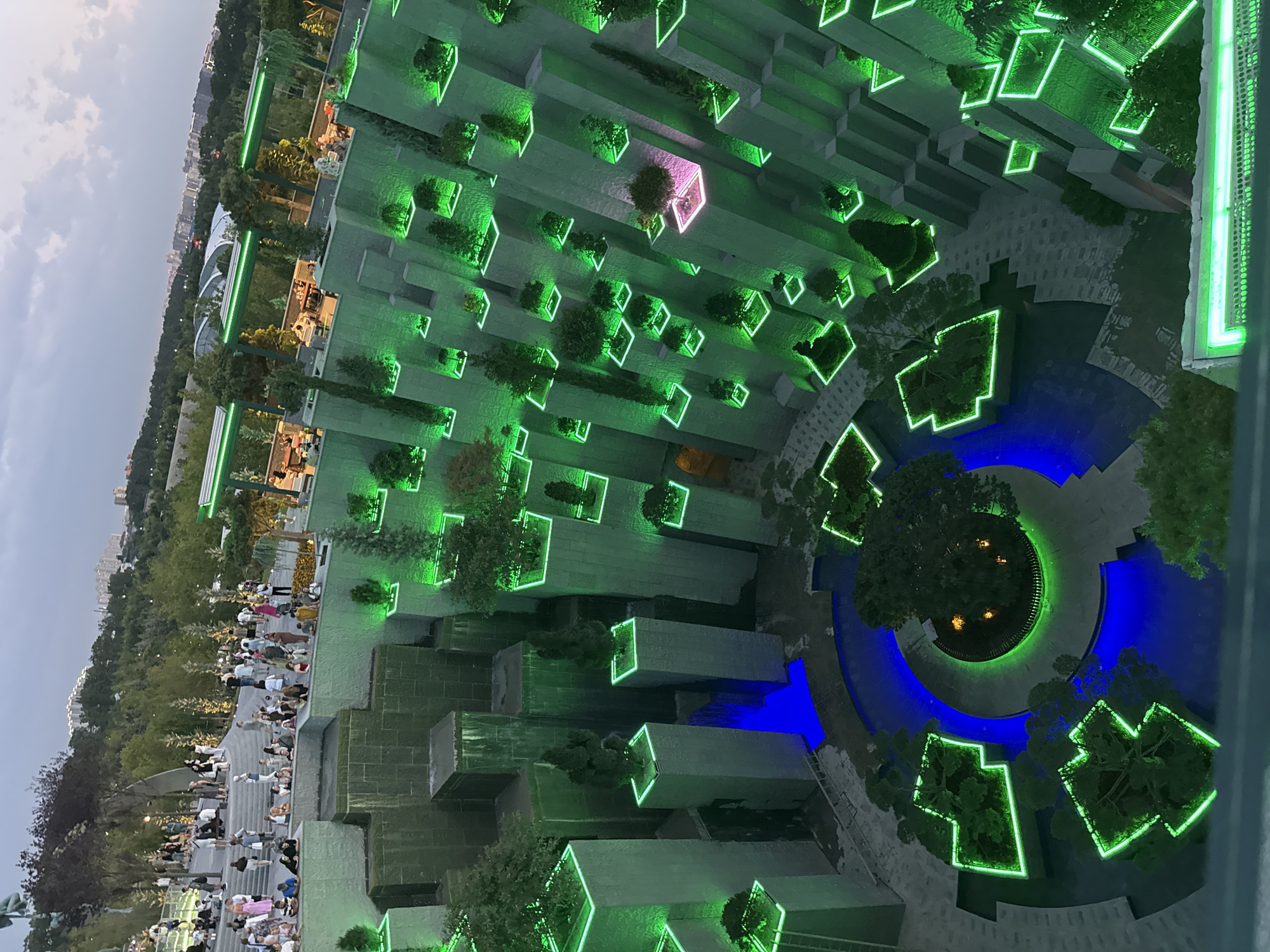 This is an elevated, wide-angle shot of a tiered, modern park or garden space at dusk, likely in Krasnodar, Russia, given the context. The architecture features stacked, geometric concrete structures with numerous plant beds and cascading water features. These elements are dramatically illuminated with vibrant green neon lights, creating a striking visual contrast with the darkening sky. Some areas of the water are lit with blue neon.
A large number of people are visible throughout the park, dispersed across various levels. On the upper terraces, a crowd of individuals is seen walking, standing, and sitting, some in groups and others individually. On lower levels, people are observed sitting at tables within covered, illuminated structures, suggesting a dining or lounging area. The overall atmosphere appears to be one of leisure and social gathering. The background reveals a cityscape with buildings and trees, indicating an urban setting. The foreground includes a railing with green neon lighting, suggesting the vantage point is from a balcony or observation deck.