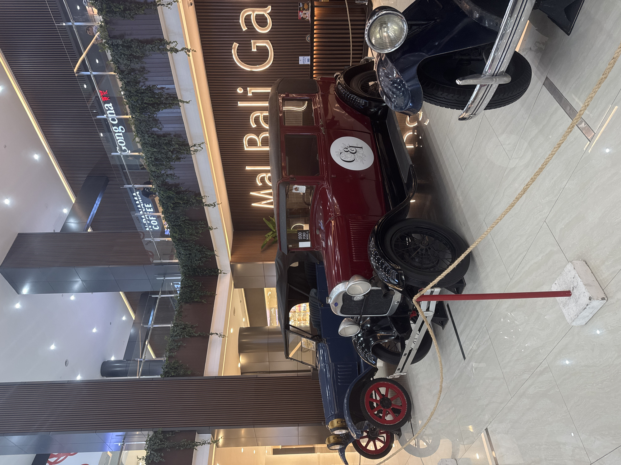 The image shows a display of vintage cars in a mall setting. In the background is a sign that reads "Mal Bali Galeria". The mall has modern architecture with wood paneling and greenery adorning the interior. The lighting is bright, suggesting an indoor environment. Two vintage cars are on display behind a rope barrier. The first is a maroon-colored car with a white emblem on the side, while the second is a navy blue-colored car. The flooring is a light-colored tile.