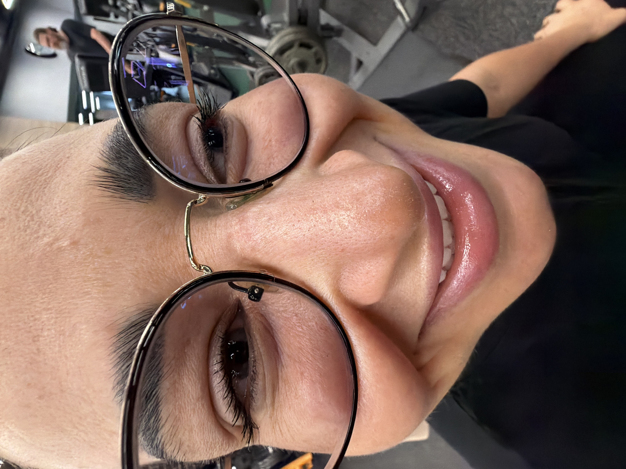 A close-up, tilted perspective photograph depicts a person with light skin wearing black-rimmed eyeglasses, smiling and positioned indoors. The eyeglasses reflect a gym setting, including another person and workout equipment, potentially indicating this photograph was taken in the city of Saint Petersburg, Russia.