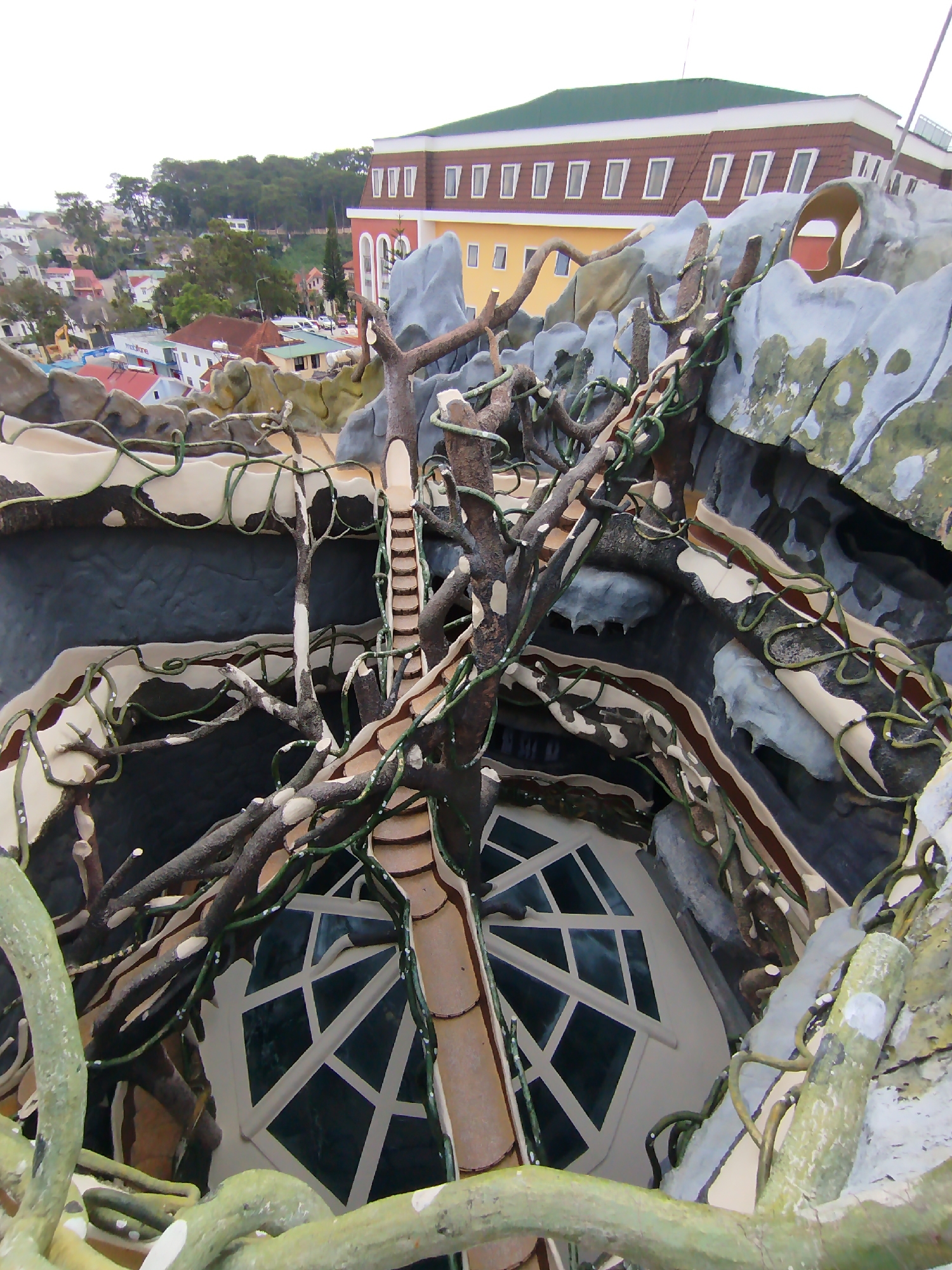 The image shows an elevated perspective of a section of the Crazy House in Da Lat, Vietnam, with a focus on a unique architectural feature resembling a tree. The tree structure has branches that connect to different levels and a staircase-like path built along one of the trunks. Below, a room with a geometric window pattern is visible. The surrounding structure is painted with various textures to mimic natural formations, with rock-like surfaces and vines incorporated into the design. In the background, city buildings with a mix of colors and roof types are seen. The sky is overcast.