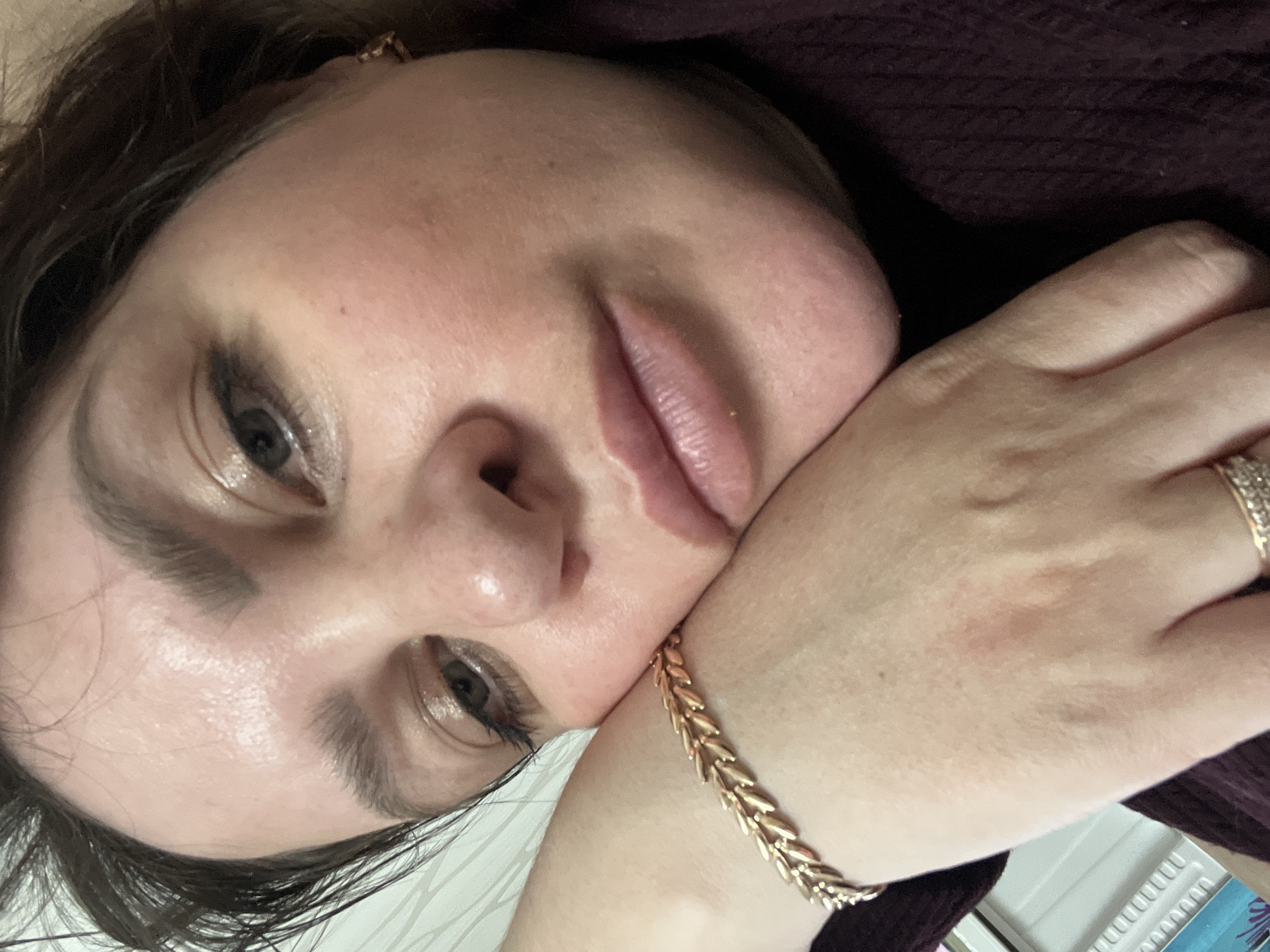 The original image presents a close-up portrait of a woman, with her head tilted and resting on her right hand. Her gaze is directed towards the viewer, conveying a calm and contemplative expression, accompanied by a subtle, gentle smile on her lips. She has fair skin with a soft rosy tone on her cheeks, and her dark brown hair partially frames her face. Her eyes feature dark eyeliner and shimmering eyeshadow, complementing her light pink or nude-toned lips. A natural sheen is visible on her forehead and nose.
On her right wrist, she wears a distinctive golden bracelet with a braided or wheat chain design, and her ring finger bears a silver-toned ring adorned with multiple small, clear stones. A slight reddish mark is discernible on the back of her hand. She is dressed in a dark, possibly burgundy or deep purple, knitted garment, visible around her shoulder and arm. The background is soft and out of focus, suggesting an indoor environment. Given the provided context, the location is Perm, Russia. There is no visible text in the image. The crops emphasize various details, including the texture of her skin, the makeup around her eyes, her lips, and the jewelry, all consistent with the main image.