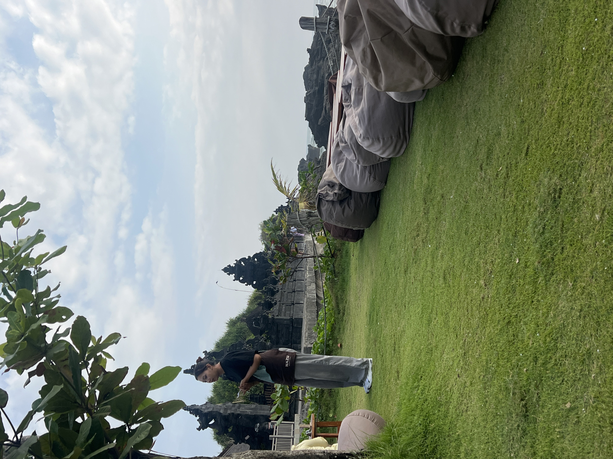 In Mengwi, Indonesia, a young woman stands on a manicured lawn in front of traditional Balinese temples and a rocky coastline. She is wearing a black t-shirt, grey pants, and white sneakers, holding a small object in her hands. To her right, a row of large, earth-toned beanbag chairs are arranged on the grass, facing the ocean. The sky above is partially cloudy, with the sun illuminating the scene from the left. The image captures a serene moment at a coastal location, likely a tourist destination or a place of spiritual significance.