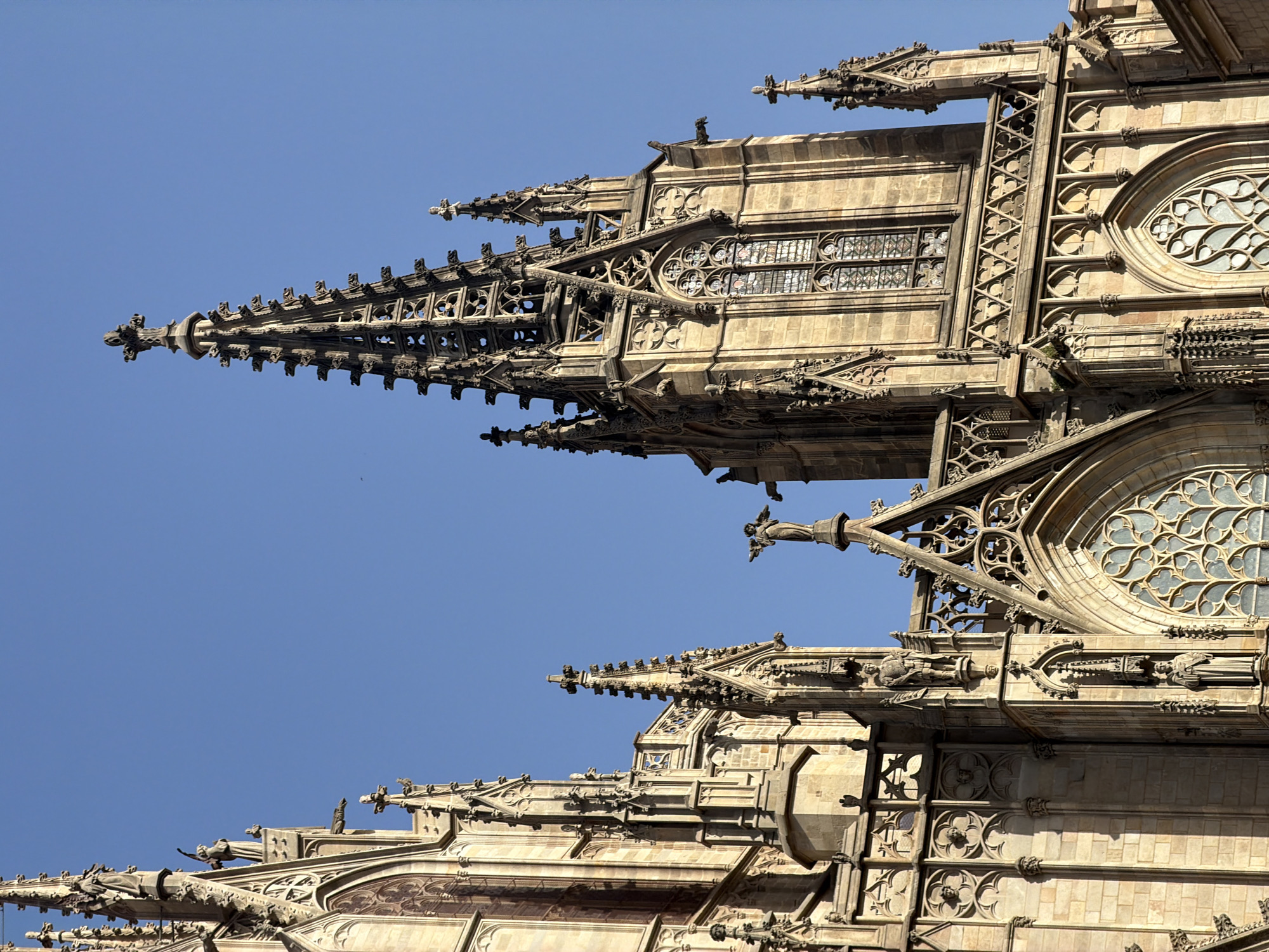 The photograph captures a close-up view of the Gothic Quarter Cathedral in Barcelona, Spain, under a clear blue sky. The image focuses on the intricate stone architecture of the cathedral's towers and spires. Numerous Gothic architectural elements are visible, including pointed arches, elaborate tracery in the windows, and detailed sculptures of saints and gargoyles adorning the facades. The stone is weathered and warm in color, illuminated by the sun, casting shadows that highlight the depth and texture of the carvings. The composition emphasizes the verticality of the structure, drawing the viewer's eye upwards towards the sky.