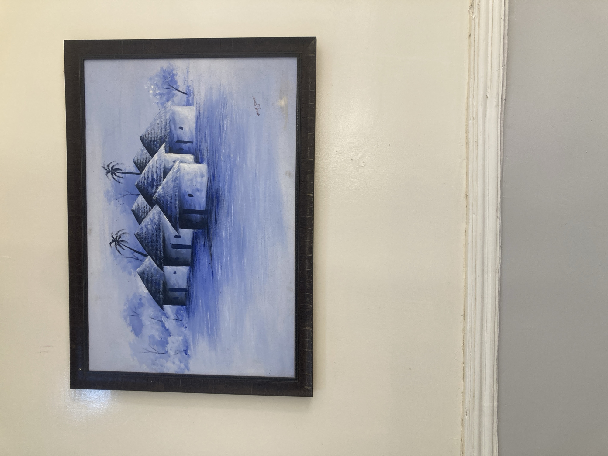 The image shows a framed painting hanging on a pale cream-colored wall. The painting is predominantly blue and depicts a waterscape with huts and palm trees. The frame is dark brown with a textured finish. To the right of the painting, the wall is divided vertically by decorative molding, with pale cream on the left and light gray on the right. No people are visible. Gura Top, Nigeria, is not mentioned in the image and cannot be confirmed as the location.