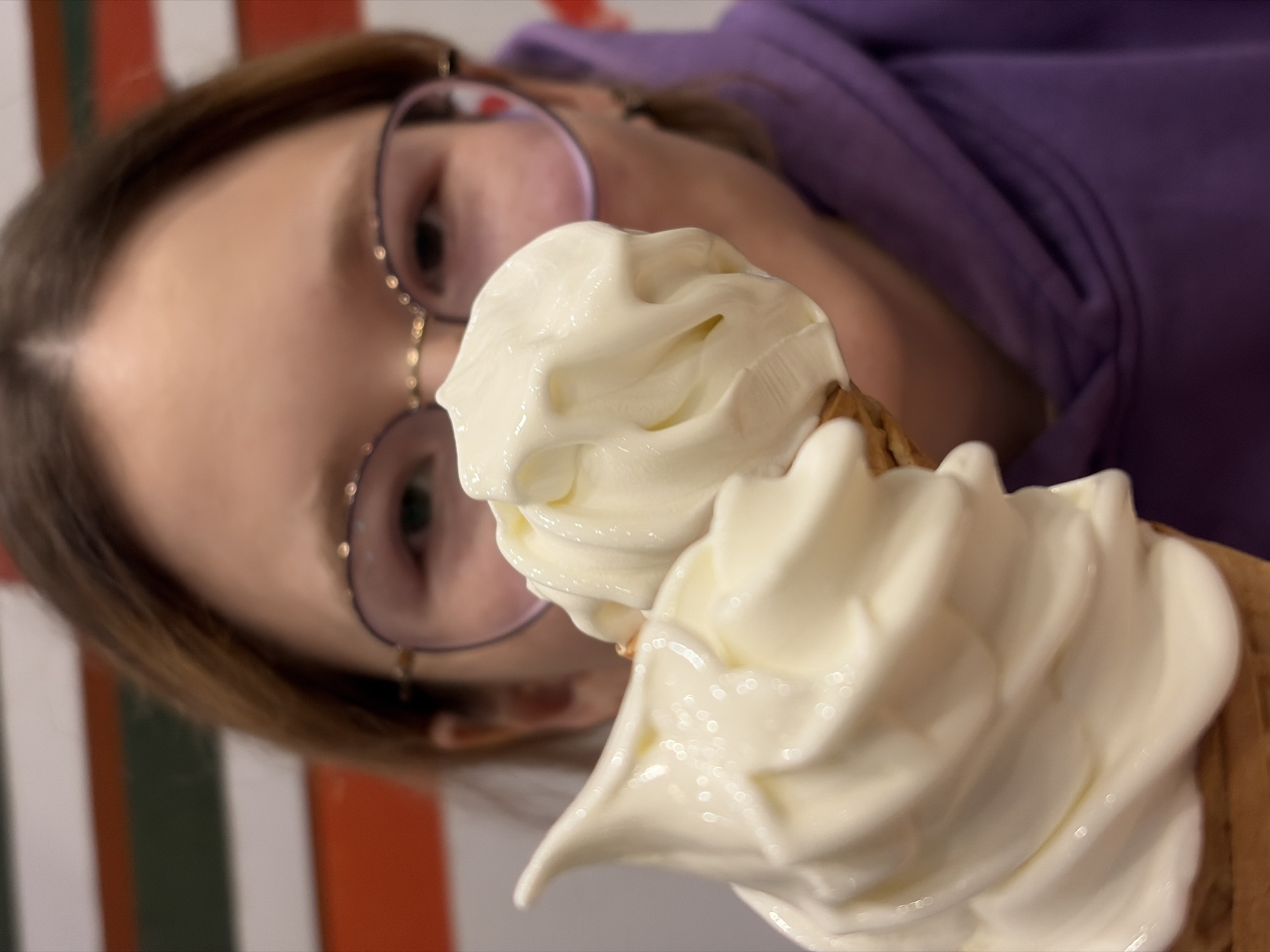 A close-up, eye-level shot shows a person wearing round, pink-rimmed glasses and a purple hoodie, holding a vanilla soft-serve ice cream cone in front of their face. The ice cream is the primary focus, with its swirls and creamy texture prominently displayed. The person's face is slightly out of focus behind the ice cream, but their eyes are visible through the glasses. The background is blurred, with horizontal stripes of red, white, and green. The location is described as Unknown, Unknown.