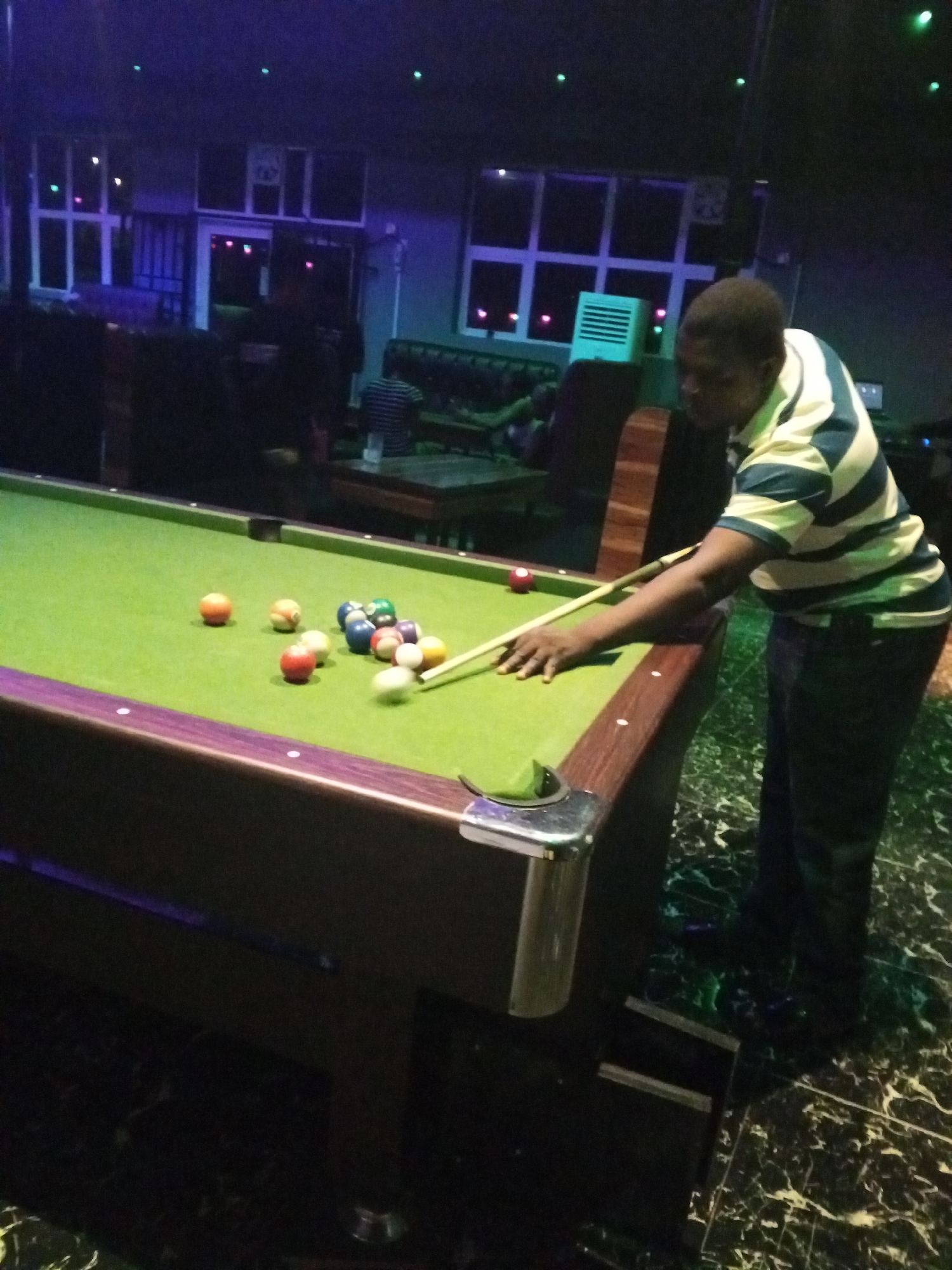 An individual is shown playing pool in what appears to be a dimly lit recreational establishment. The person, wearing a striped shirt and dark pants, is positioned at a pool table covered in green felt, with several billiard balls arranged on it. The pool cue is held, suggesting the person is in the act of taking a shot. In the background, there are tables and chairs, and other individuals can be seen in the dimly lit area. The environment is suggestive of a bar or pub, though the specific location is unknown. The pool table has a dark wood trim with metal corner protectors. The floor has a shiny black and green marbled design.