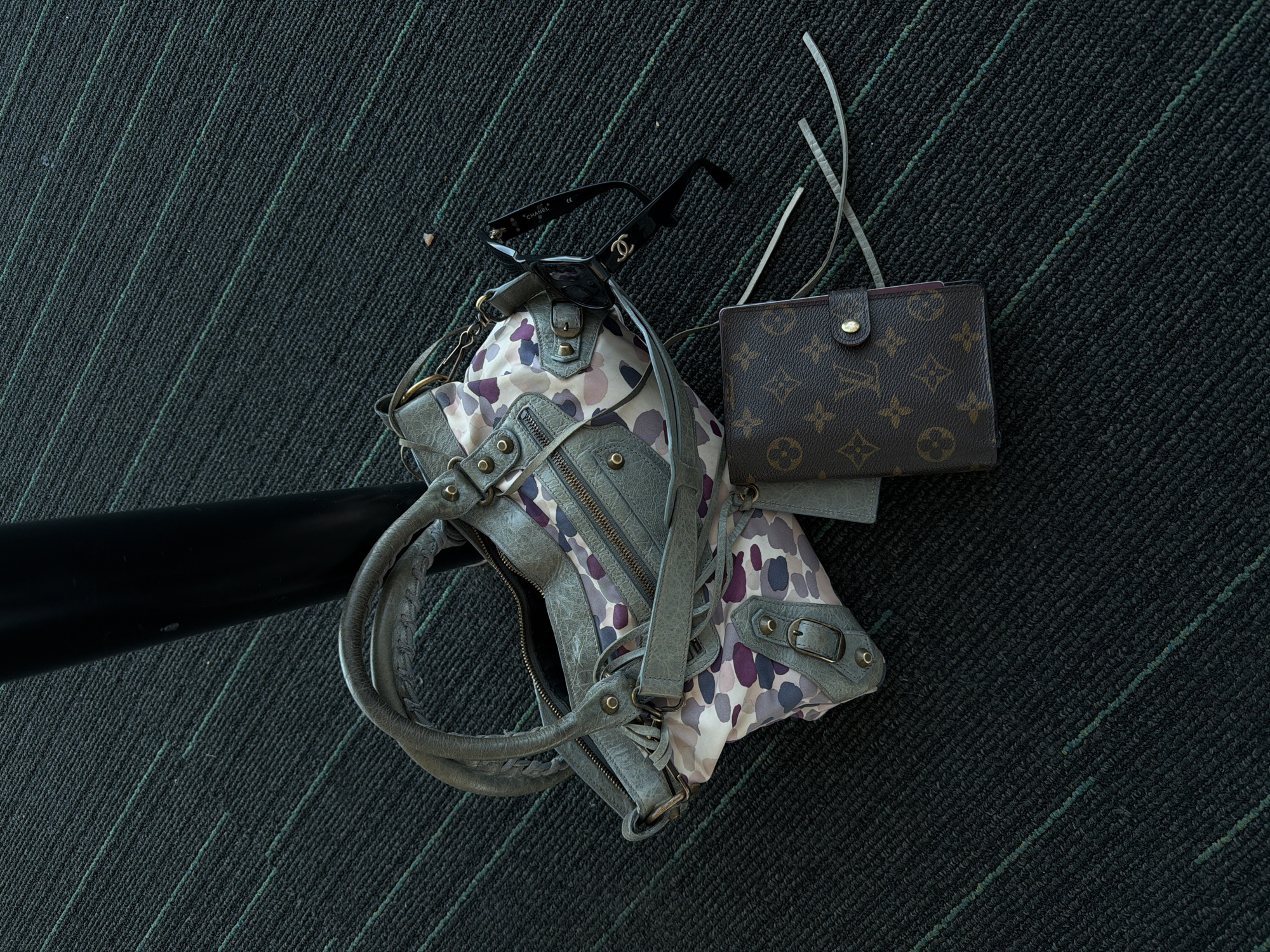 The image depicts a still life arrangement of personal belongings against a dark carpet with subtle green linear patterns. A handbag, predominantly gray with a printed fabric accent featuring purple and white blotches, lies open, revealing its interior. Atop the handbag rests a pair of black Chanel sunglasses. To the right of the handbag, a brown Louis Vuitton wallet is positioned. The background is dominated by the dark carpet, which appears to be tightly woven. A portion of a black, cylindrical object, possibly a bar or pole, is visible towards the left side of the frame. The scene appears to be indoors. There is no information to make a determination about the location in Uttlesford, UK.