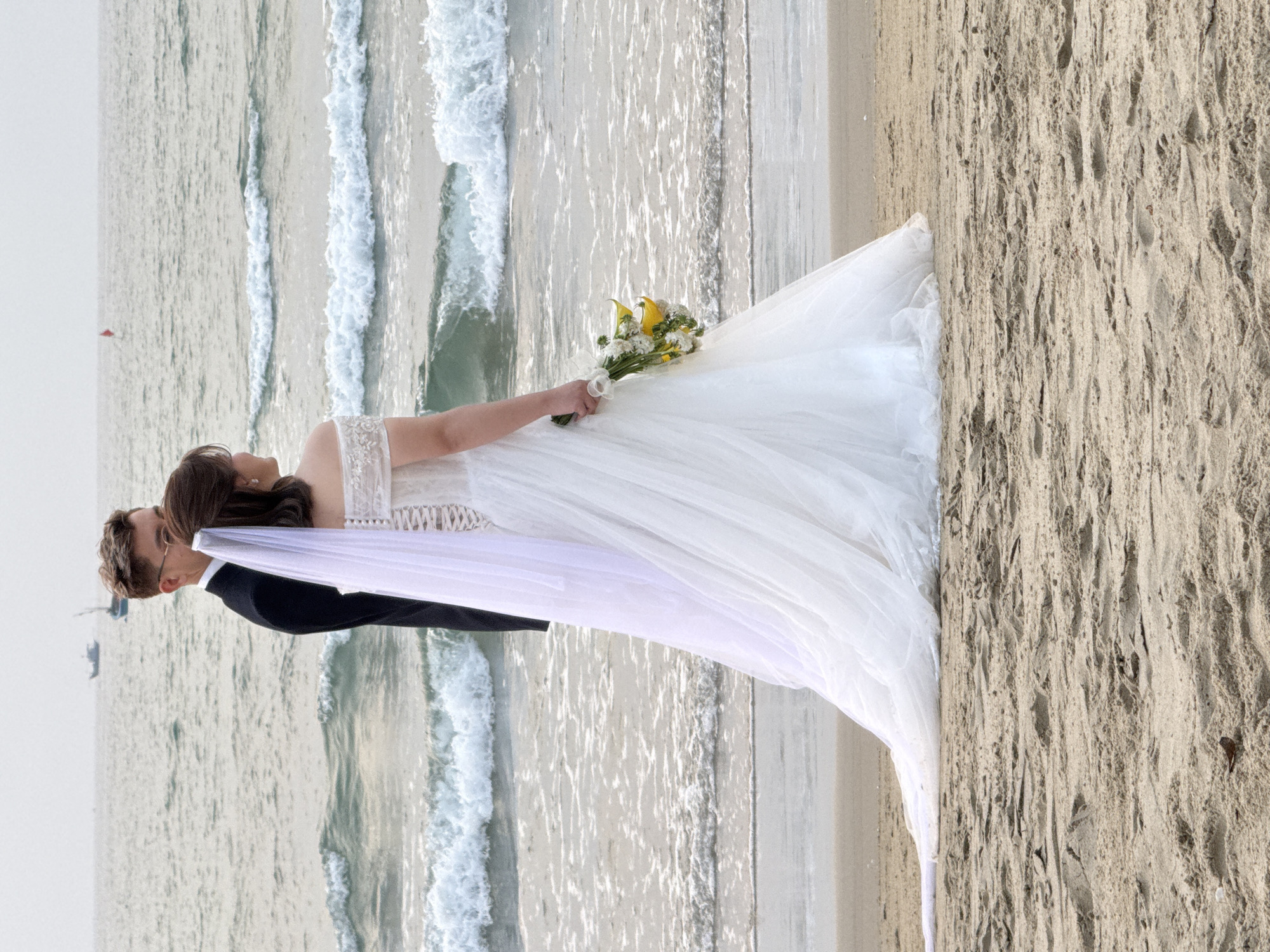 A wedding photo shows a bride and groom on a beach. The groom, wearing a black suit, stands close to the bride, whose face is partially obscured by her dark hair. The bride wears a white wedding dress and holds a bouquet of yellow and white flowers. A white veil extends from her head. Behind them are waves of the ocean. The scene also shows the sandy beach and sky. While the specific location within Đà Nẵng, Vietnam, cannot be pinpointed from visual data alone, the beach setting is consistent with the coastal geography of that city.