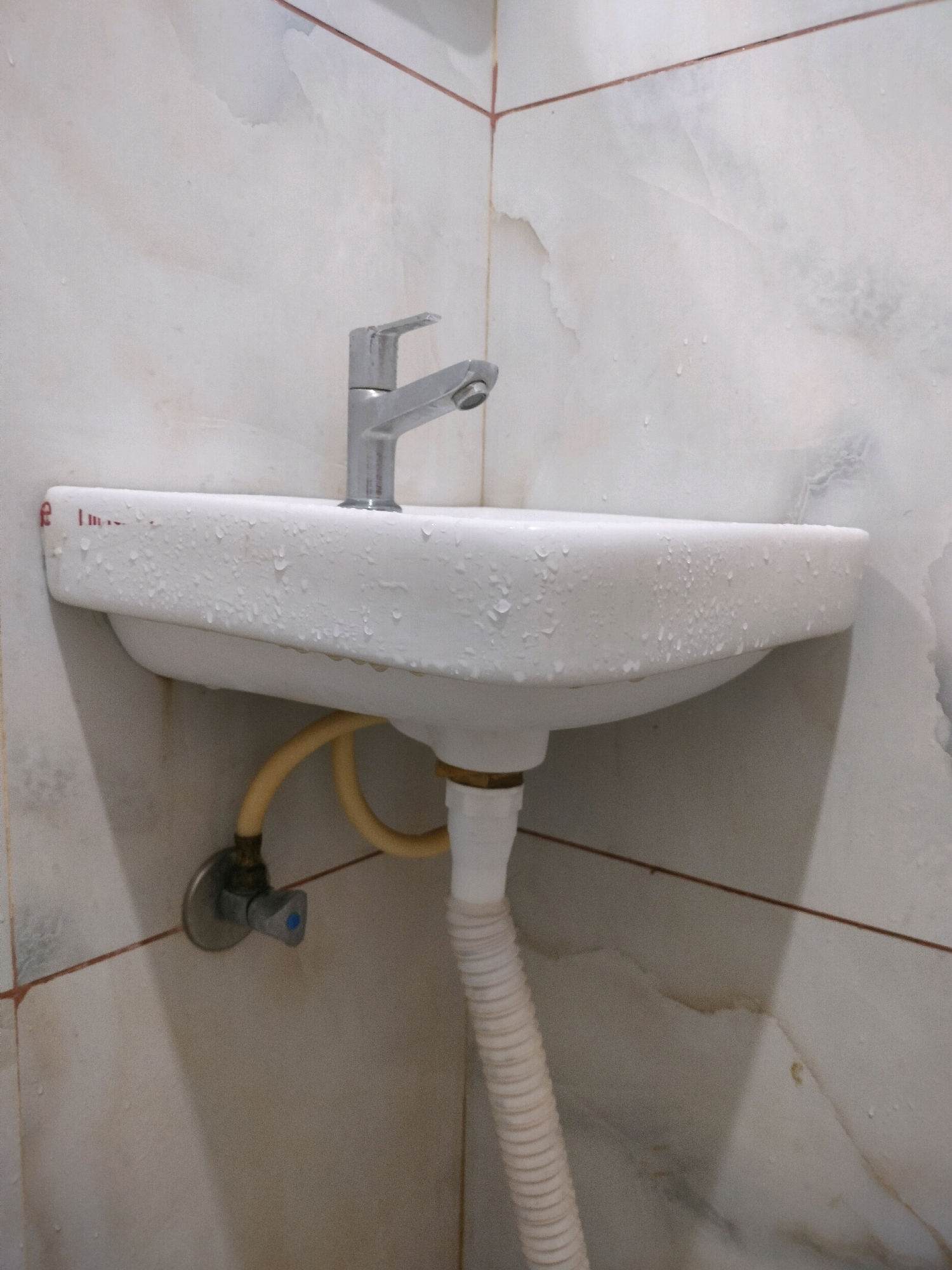 A white ceramic sink is mounted on a tiled wall in what appears to be a bathroom. A silver faucet is attached to the sink. Water droplets are visible on the surface of the sink and the faucet, suggesting recent use or a leak. Below the sink, a flexible yellow hose connects to a plumbing fixture, and a white corrugated pipe extends downwards. The wall tiles are a light color with visible grout lines and some water stains. The scene is dimly lit. The presence of a sink with plumbing suggests an indoor setting, likely a residential or public restroom. This image does not contain enough information to determine its specific location within Bengaluru, India, or to identify any people or specific actions.