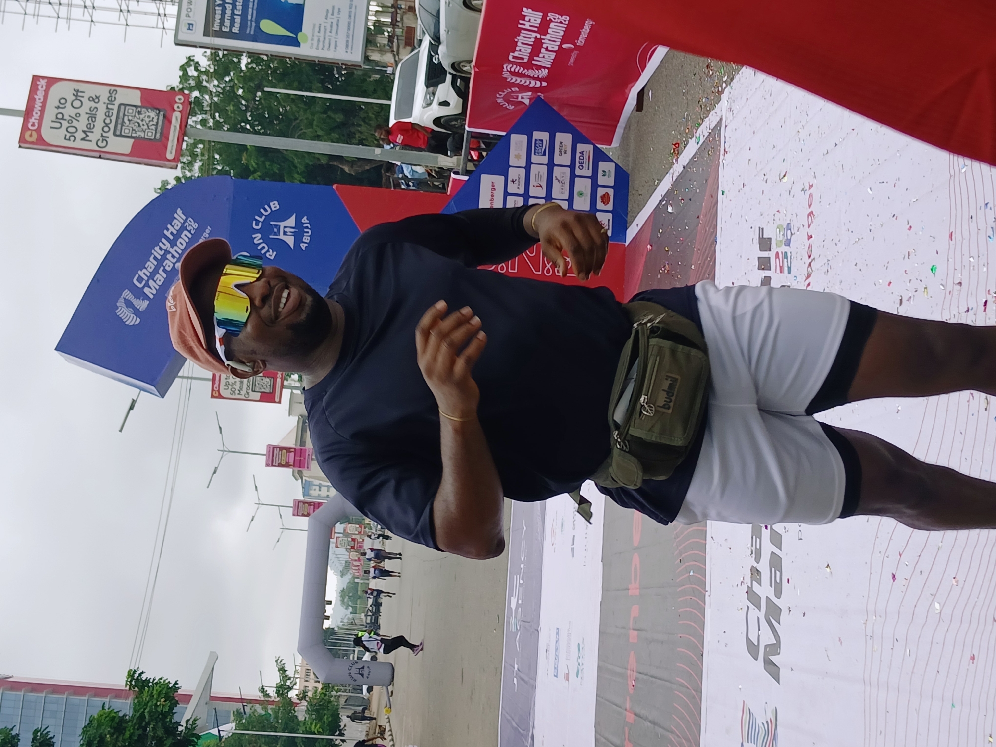 A man wearing a dark blue t-shirt, white shorts with black trim, a green fanny pack, and distinctive iridescent sunglasses is smiling and looking towards the left. He is standing on a white tarp with confetti scattered on it, possibly indicating the finish line of a race. Behind him, an inflatable archway marks the start or finish of a "Charity Half Marathon 2023" hosted by "RUN CLUB ABUJA." Multiple runners are visible behind the archway, moving forward. In the background, there are buildings, traffic, streetlights, and various signs, including one for "Chowdeck" offering discounts on meals and groceries, and another for "Power Stone Real Estate." The sky is overcast. This scene is taking place outdoors in the city of Abuja, Nigeria.
