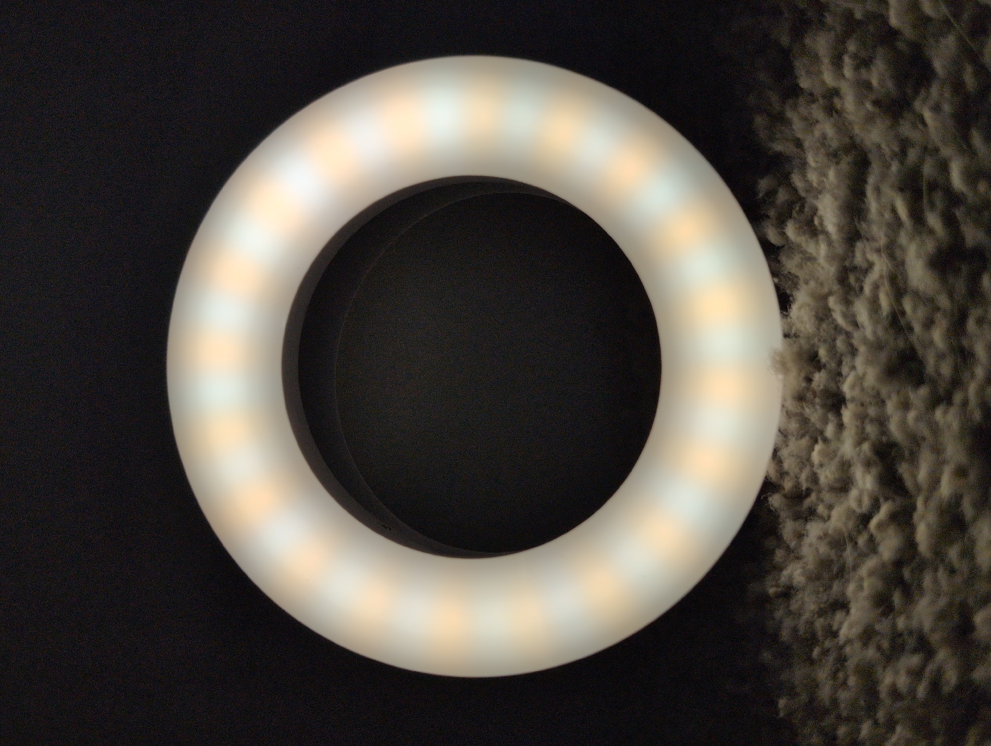 The image shows a close-up, high-angle shot of a white ring light with yellow and white LED lights, placed on a dark, fuzzy surface that appears to be carpet or fabric. The ring light is the primary subject, positioned in the center of the frame. The background is dark and out of focus. The lighting from the ring light casts a glow, creating a circular illumination effect. The overall scene is static and focuses on the object and its immediate surroundings. There are no people or discernible landmarks of Louisville, United States of America visible in the image.