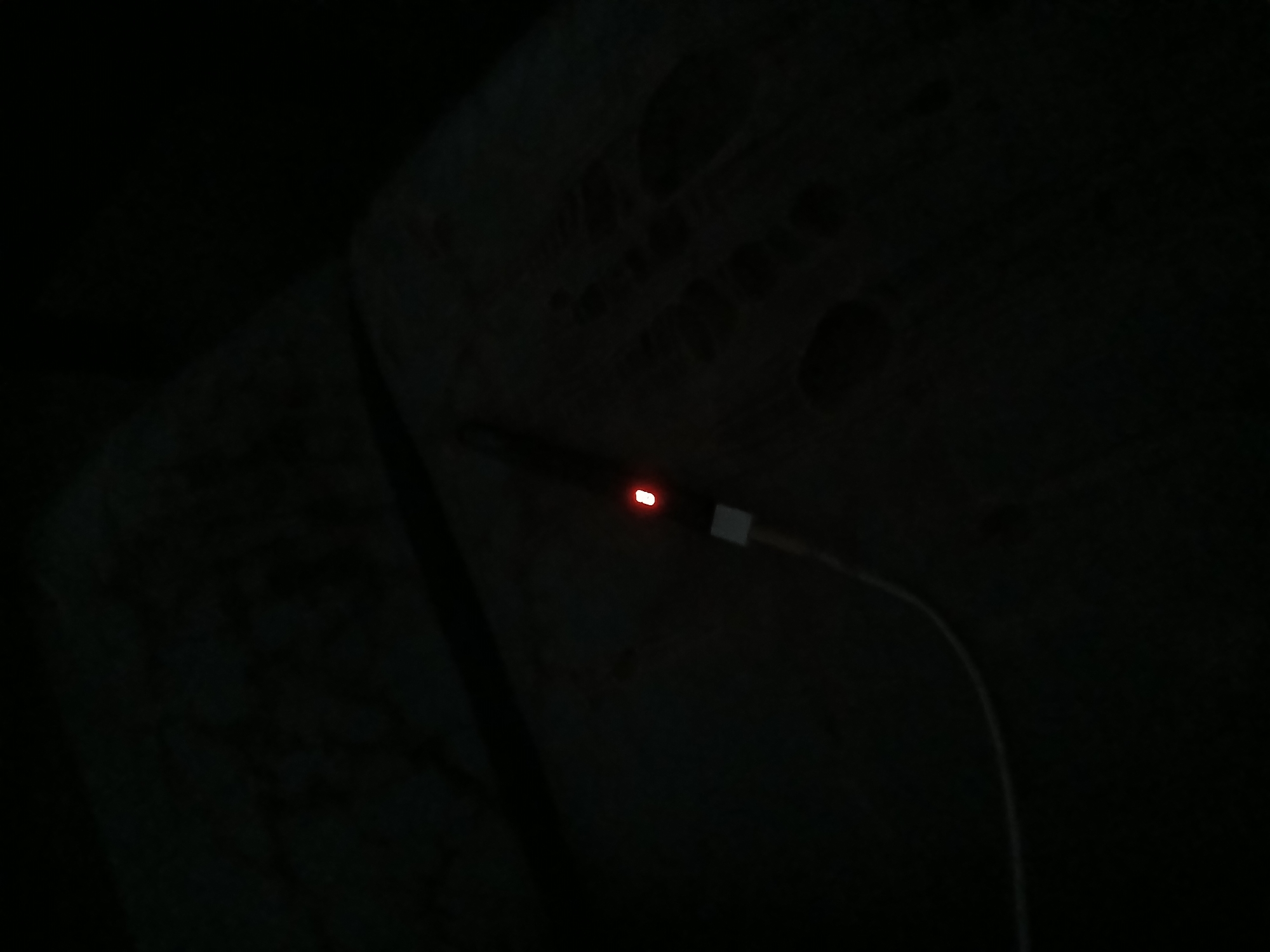 In a dark environment, a white USB cable extends from the bottom left corner towards the center of the frame. The cable connects to a small white adapter which is attached to a dark electronic device. A small red LED light is illuminated on the device, suggesting it is powered on or charging. The background is dark and indistinct, but appears to be a textured surface. The scene appears to be indoors, possibly a home or office setting in Kademangan Kulon, Indonesia.