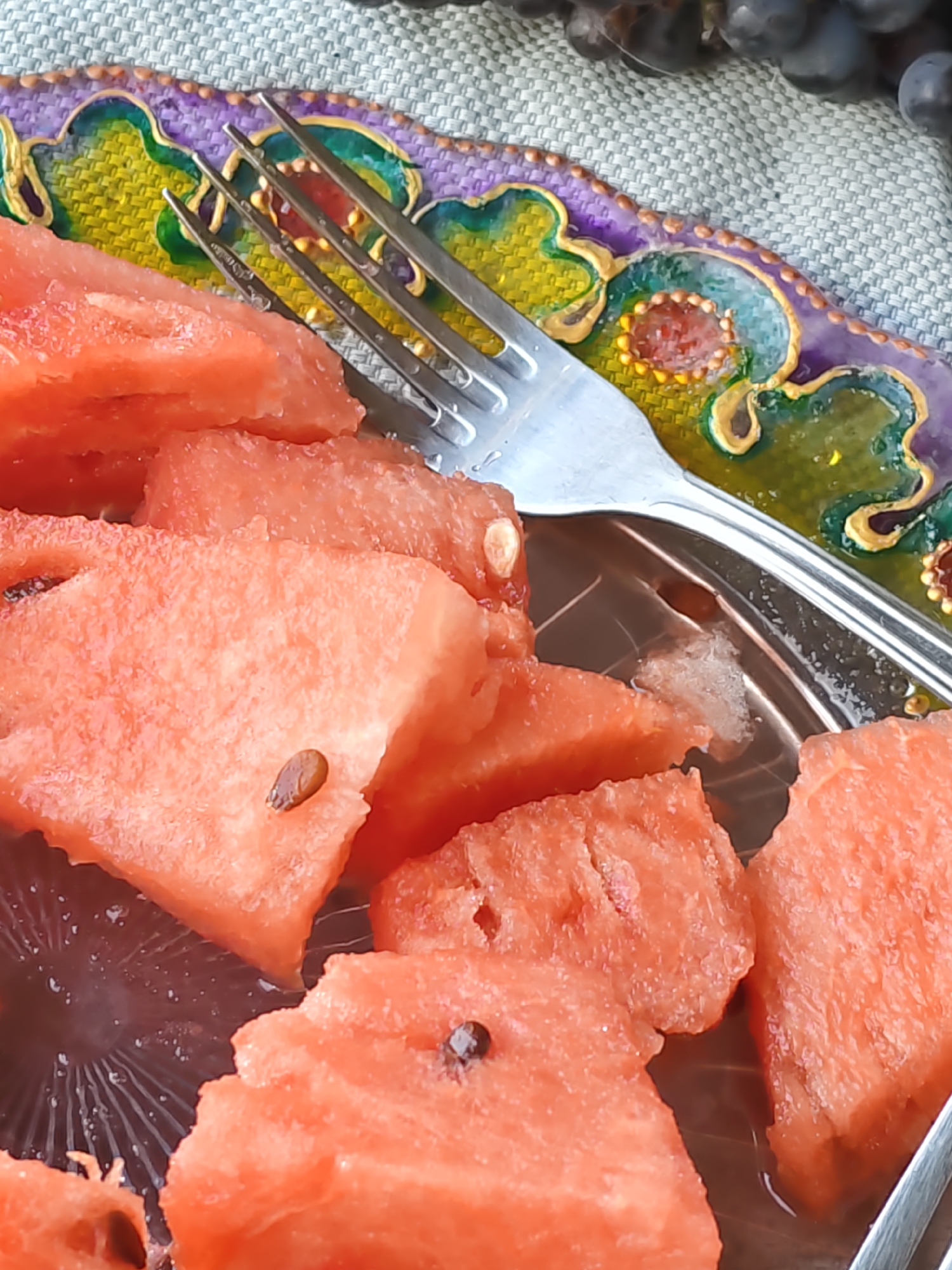 The image shows a close-up of several sliced pieces of watermelon arranged on a decorative platter. A silver fork is resting on top of some of the watermelon slices. The platter has a colorful, ornate rim with purple, green, and gold detailing. The watermelon pieces are vibrant pink and some seeds are visible. In the upper right background, a cluster of dark grapes can be seen, resting on a textured grey surface, possibly a tablecloth or placemat. There is no indication of the city of Rostov-on-Don, Russia, in the image itself.