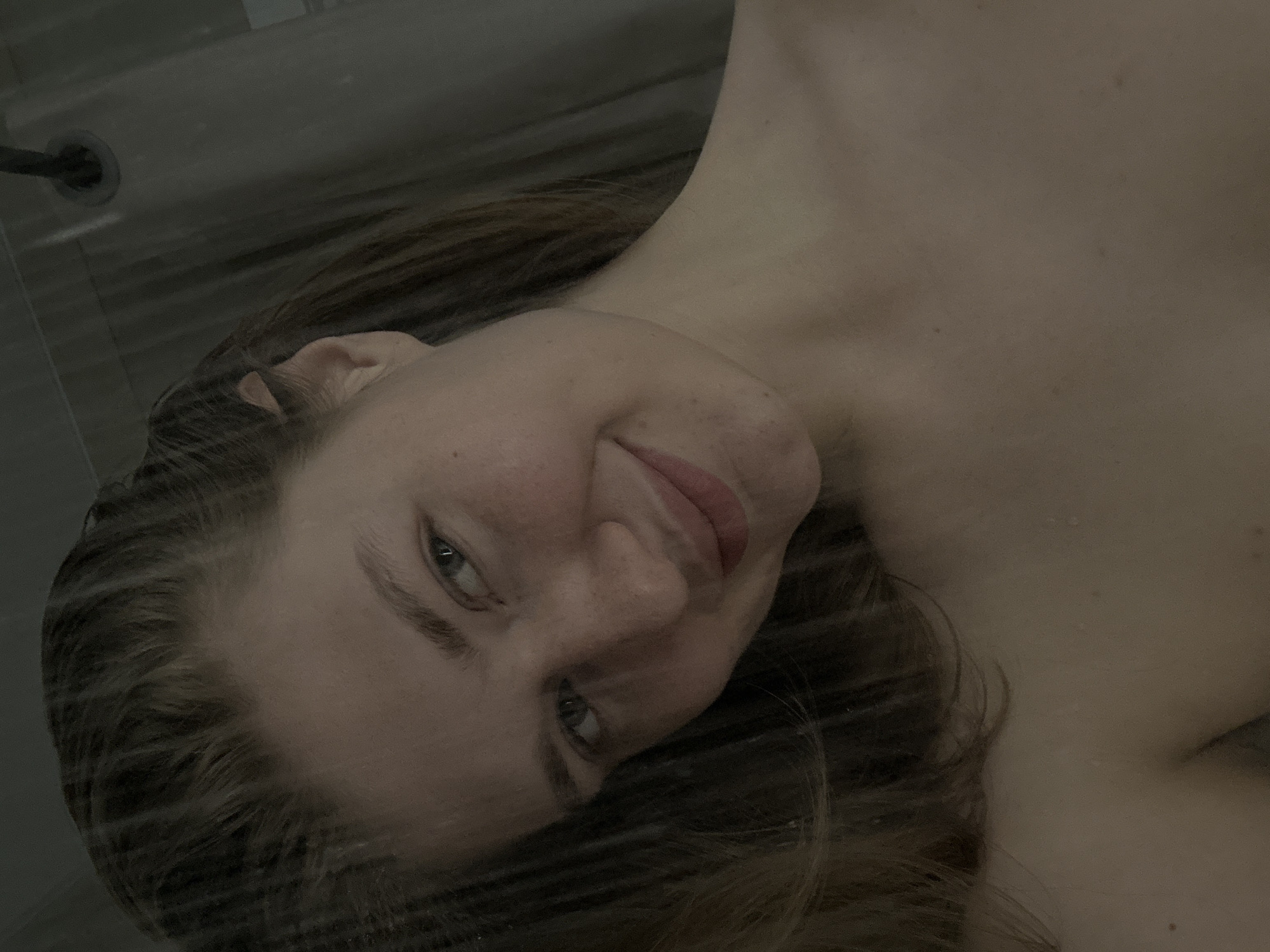 A close-up, slightly angled shot depicts a person with light skin and brown hair. The individual is partially obscured by water droplets, suggesting they are in a shower. A showerhead is visible in the upper left corner of the frame. The location is likely a private bathroom in an unknown city.