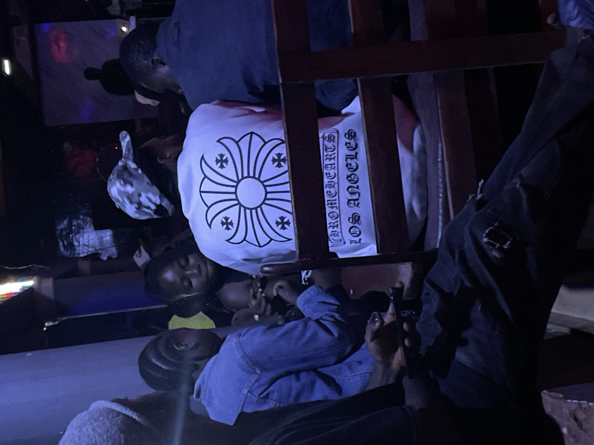 The image depicts a dimly lit indoor scene, likely a social gathering or club in Durumi, Nigeria. Several individuals are visible, seated and facing away from the camera. In the foreground, a person wearing a white t-shirt with a distinctive floral and cross design, along with the text "CHROMEHEARTS LOS ANGELES," is prominent. To their left, another individual wears a denim jacket. Further left, a person with a grey beanie is partially visible. To the right of the person in the white t-shirt, another individual in a dark shirt is seated. The environment appears to be a public space with wooden seating and ambient lighting that casts a blue hue. The background is dark, with some obscured figures and possibly a screen or projection. The overall atmosphere suggests a casual social setting.