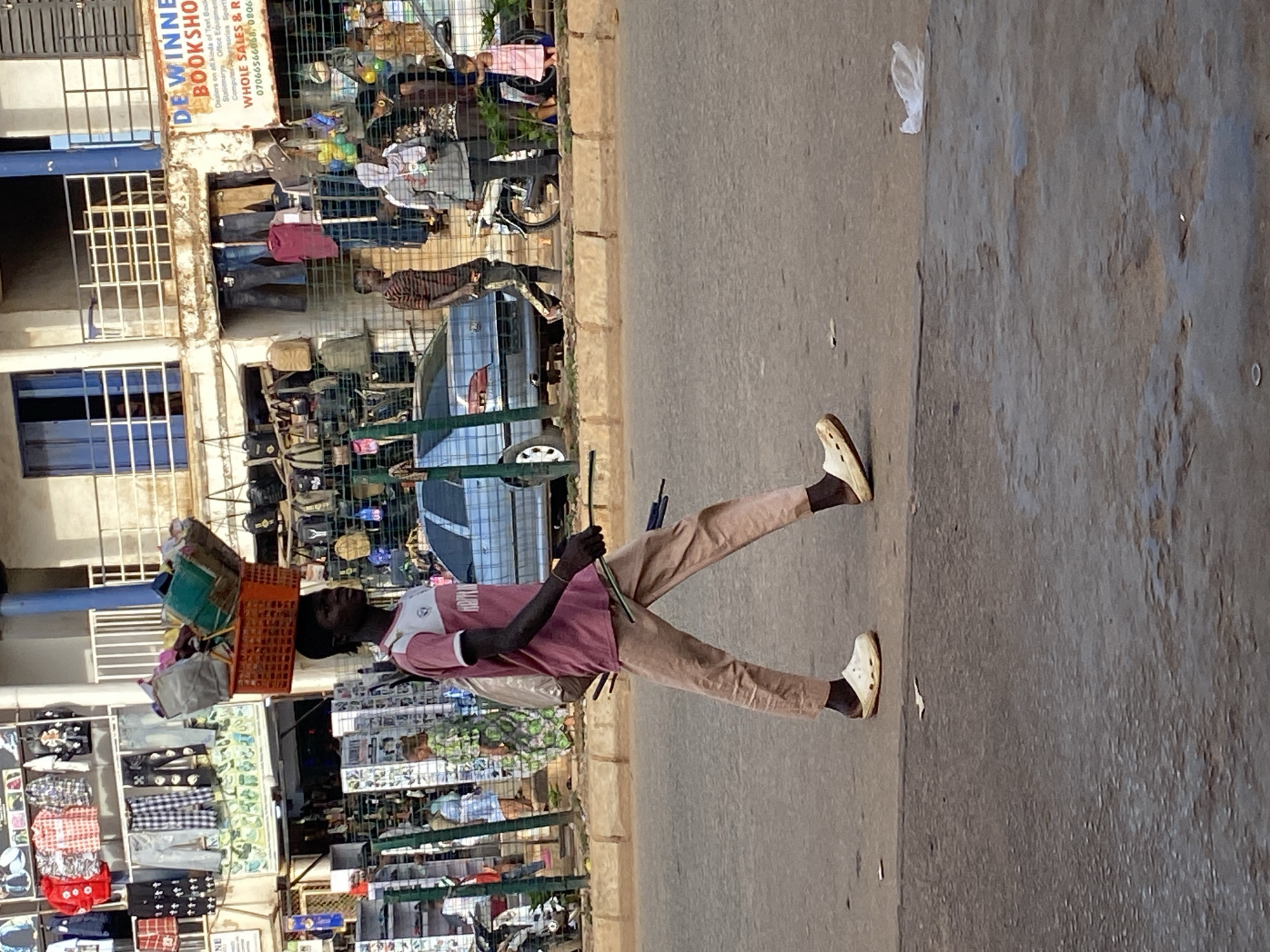 A man carries goods on his head on a city street in Bukuru, Nigeria. He is wearing a pink shirt, khaki pants, and white Crocs shoes. He carries a stick in his right hand. The goods on his head include items in a red plastic crate. He walks on a concrete street. Behind him is a building displaying goods and a bookstore with the name "DE WINNE BOOKSHOP" on the sign. A car is parked nearby.