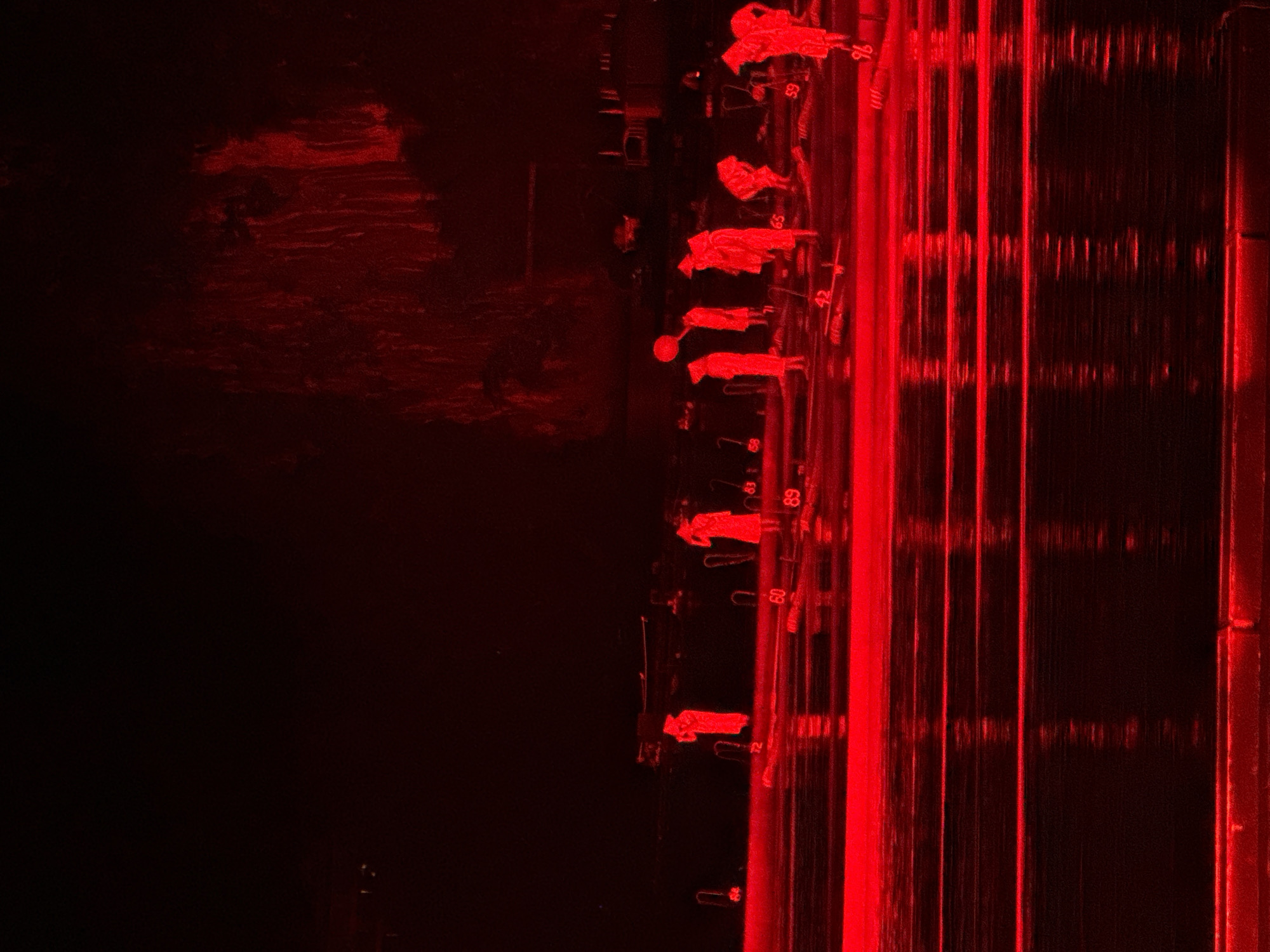 The image shows a red-lit scene with a dark area on the left and brighter, illuminated structures on the right. Along a horizontal line in the middle, there are several indistinct figures or objects, some possibly resembling people in clothing or robes, arranged in a row. Numbers are visible near some of the figures. To the right, there are parallel vertical lines or columns of light, possibly reflecting off water. The overall color scheme is red, suggesting artificial lighting or a filter effect. The location or specific details related to Guilin, China, cannot be determined from the image alone.