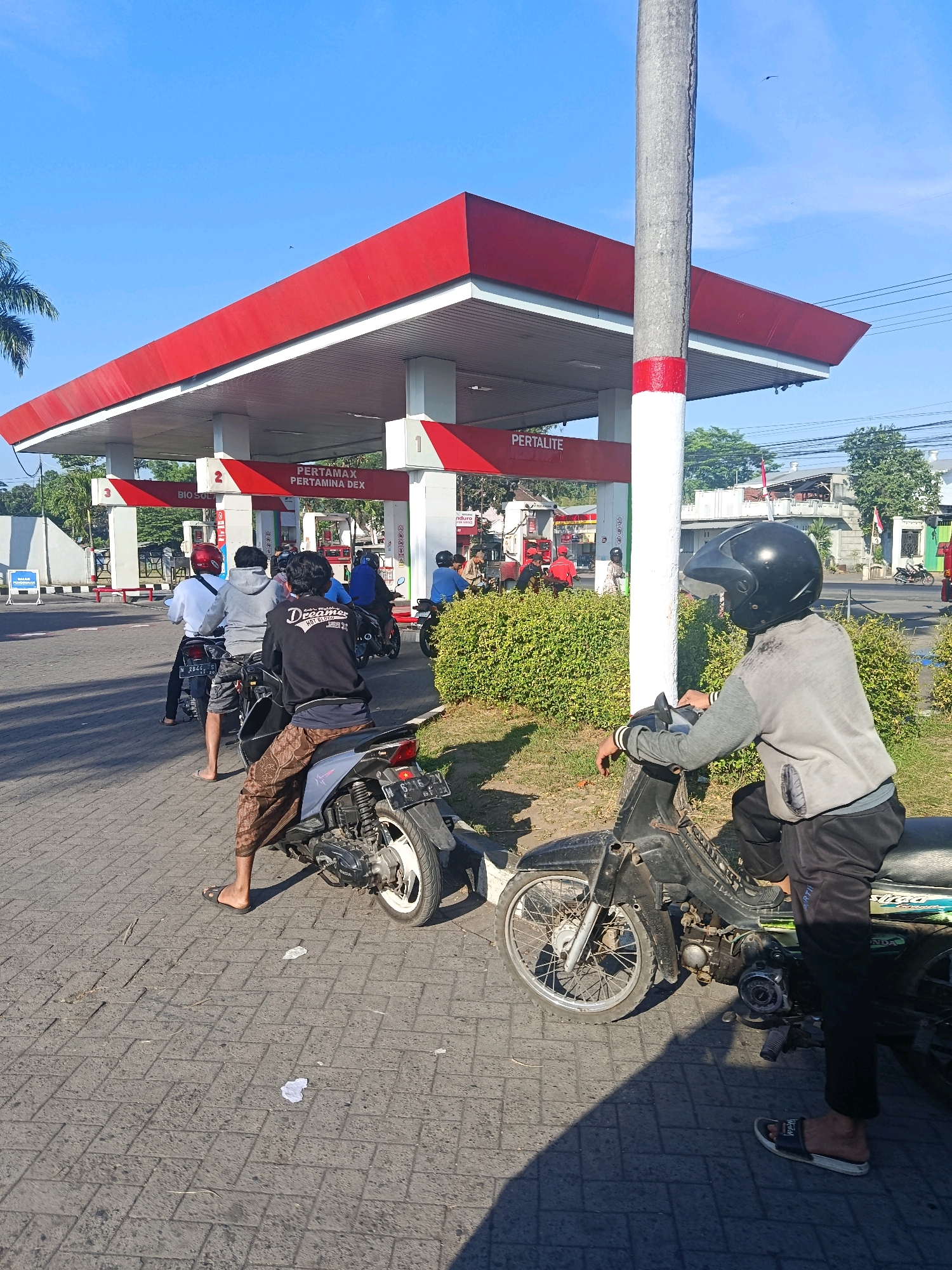 This image captures a scene at a Pertamina gas station in Tempeh Lor, Indonesia, under a clear blue sky. Several people on motorcycles are lined up, waiting to refuel. A concrete pillar, painted with red and white bands, stands prominently in the foreground. The gas station canopy is red with white supports, and signage indicates "PERTAMAX PERTAMINA DEX" and "PERTALITE." Green bushes line the side of the paved area. In the foreground, two individuals are visible on their motorcycles, one wearing a black helmet and grey jacket, and another in the background wearing a red helmet. The ground is paved with interlocking bricks. The atmosphere appears to be daytime, with the sun casting shadows.