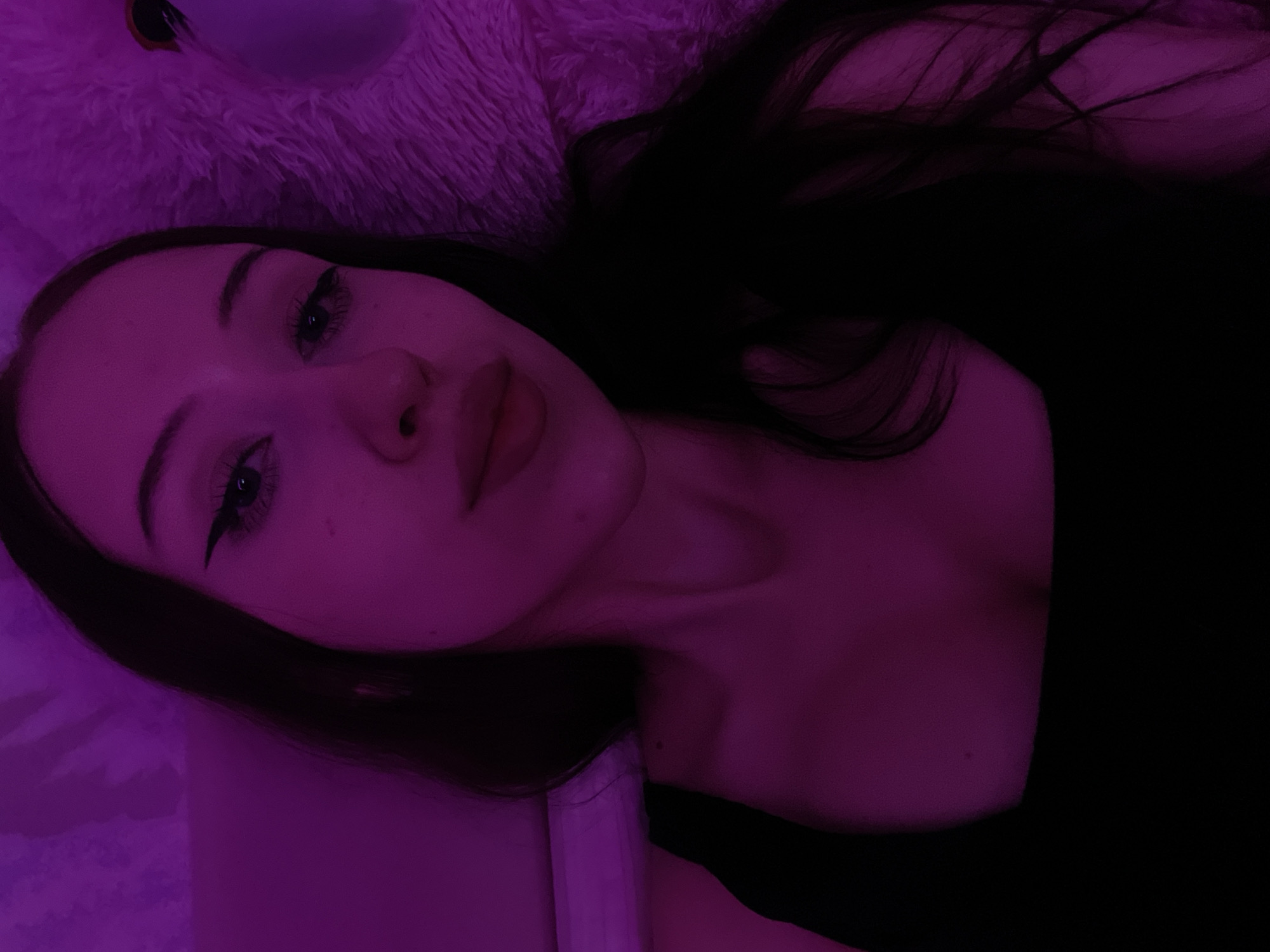 A young woman with long, dark hair poses for a selfie in a dimly lit room, illuminated by a purple light source. She is wearing a black tank top. Her makeup includes winged eyeliner and a matte lipstick. To her right, a fluffy pink object, possibly a stuffed animal or blanket, is visible. The background appears to be a wall or bedspread, also cast in the purple hue. There is no indication of the location being Votkinsk, Russia.