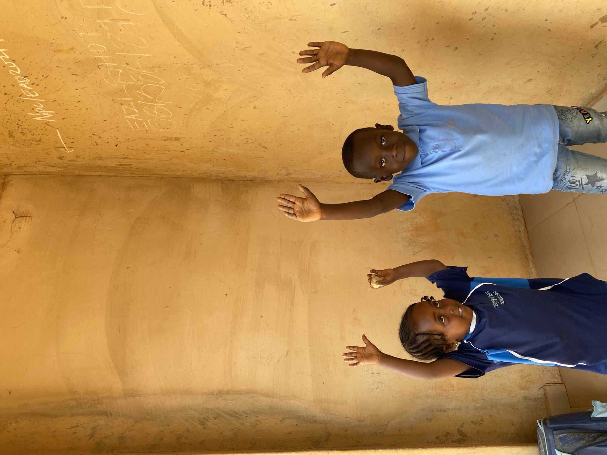 Two children pose for the camera in an indoor setting, likely a school in Kuru Dungus, Nigeria. The walls are a faded yellow, with graffiti in white chalk or similar material. A boy in a light blue collared shirt and jeans is above, while a girl in a navy blue and light blue school uniform is below. Both children have their arms raised, as if reaching towards the camera. A portion of what appears to be luggage is visible in the lower right corner.