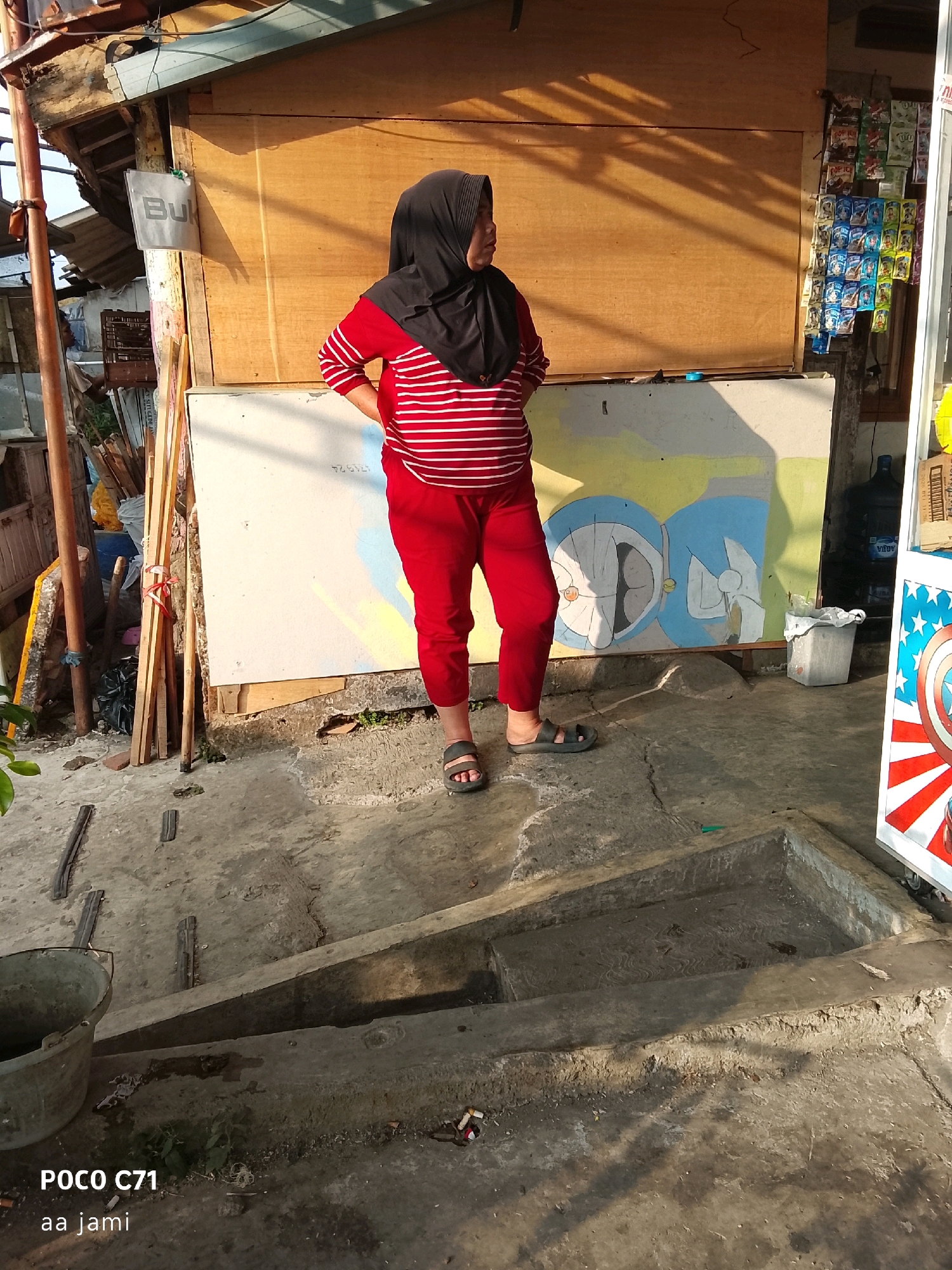 A full-body shot captures a woman from the knees up, standing outside a small outdoor shop. The woman is wearing a black hijab and a red and white striped long-sleeved shirt, paired with red pants and black sandals. Her stance is relaxed, with her left hand on her hip and her right arm slightly bent.
The shop appears to be constructed with a wooden facade and a corrugated metal roof. A sign with the word "Buk" is visible on the wooden wall. To the right of the woman, a section of the wall is adorned with a colorful mural featuring characters that resemble Doraemon, a popular Japanese anime character.
In front of the shop, the ground is concrete, with some visible debris and plants growing in cracks. A large grey bucket is situated to the left, and further to the right, a blue water dispenser is visible. The overall environment suggests a modest, possibly informal commercial setting. The lighting indicates it is daytime, with shadows cast by the shop's roof.