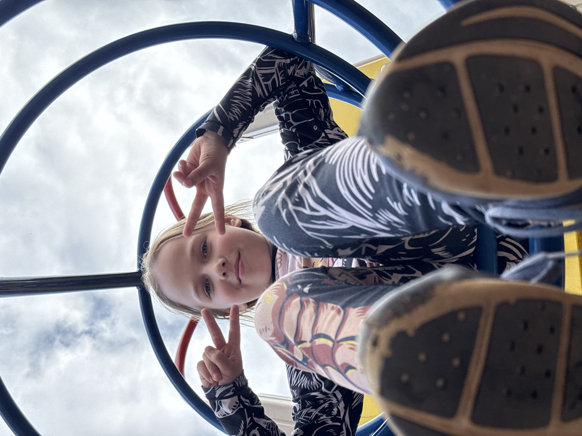 A low-angle, outdoor, close-up, fisheye shot captures a young girl from below, looking up as she poses on a playground structure. The girl, with blonde hair and blue eyes, is positioned in the center of the frame, her face partially obscured by her knees and legs. She is wearing a black and white patterned long-sleeved shirt and leggings with a similar pattern, and her legs are bent as she sits or crouches on the equipment. She makes a peace sign with both hands, holding them up near her face. The soles of her black shoes are prominently featured in the foreground, closest to the camera, with a textured pattern.
The playground structure is visible with blue and red bars forming arches around the girl. In the background, a cloudy sky is visible through the bars. The perspective suggests the photographer is lying on the ground beneath the playground equipment. The image is well-lit, indicating it was taken during the daytime, under overcast conditions. The overall mood is playful and energetic, typical of a child enjoying an outdoor activity. No visible text is present in the image.