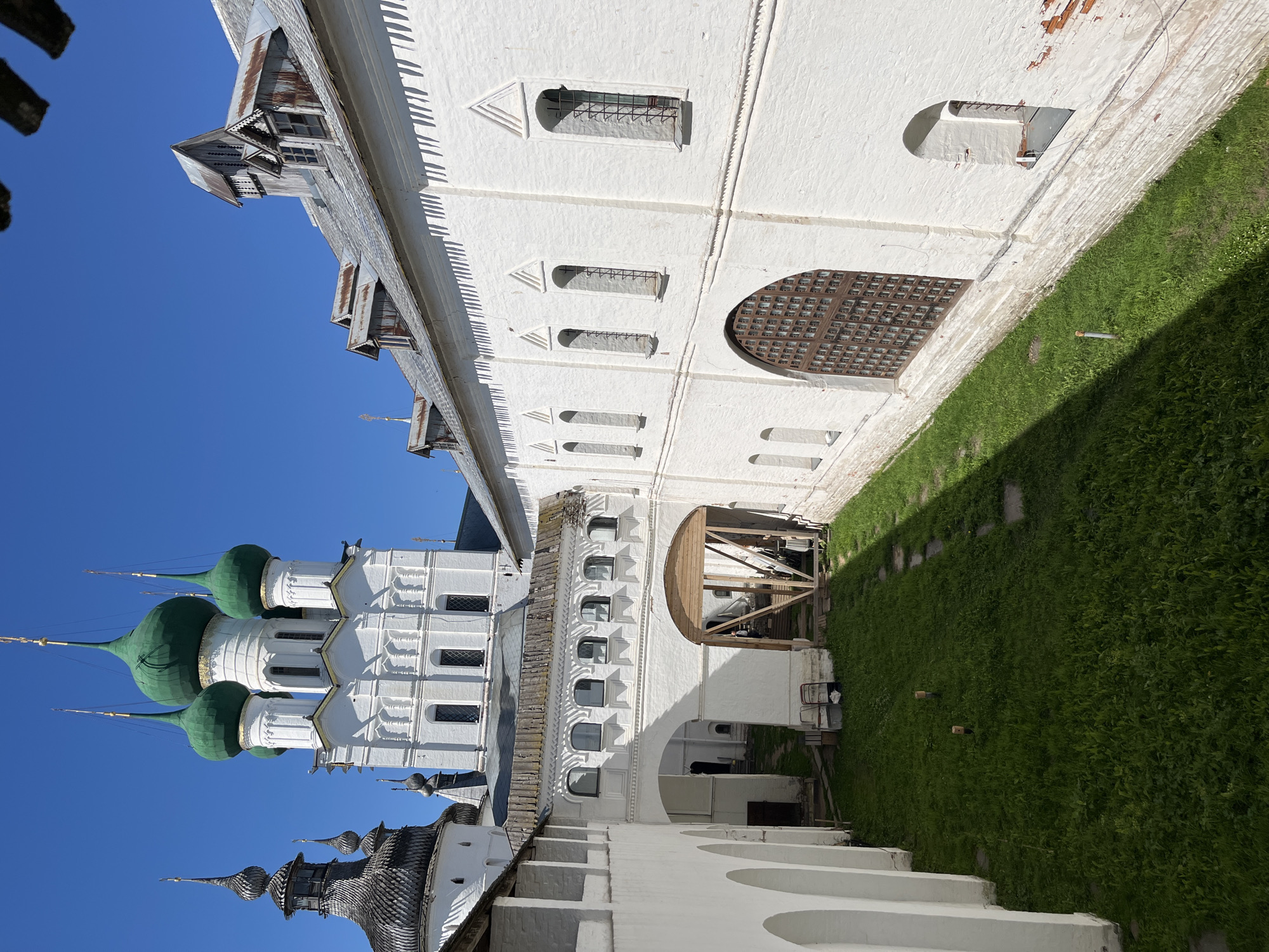 The image shows a view of the Rostov Kremlin complex in Kladovitsy, Russia. The foreground features white stone walls with arched walkways and stairs, covered in a vibrant green lawn. In the mid-ground, a white building with green-domed roofs stands against a clear blue sky. The building's architecture includes multiple windows and decorative elements. Further in the background, other structures with similar white walls and roofs are visible, including some with dark, ornate spires. The scene suggests a bright, sunny day, possibly in the late morning or early afternoon, given the clear sky and strong shadows cast by the buildings onto the grass. The overall atmosphere is serene and historical, indicative of a religious or cultural heritage site. There is no visible text in the image.