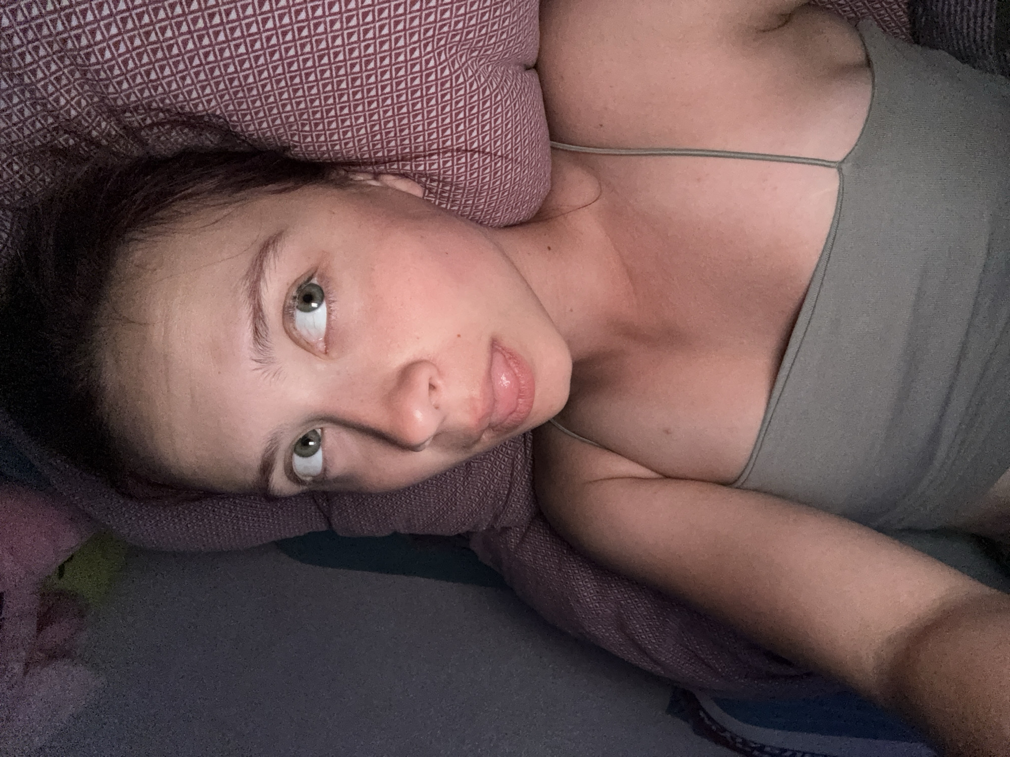 A woman is lying on a bed. Her head rests on a pillow with a patterned cover. She is wearing a light green tank top. Her eyes are open and she is looking at the camera. The location is not specified.