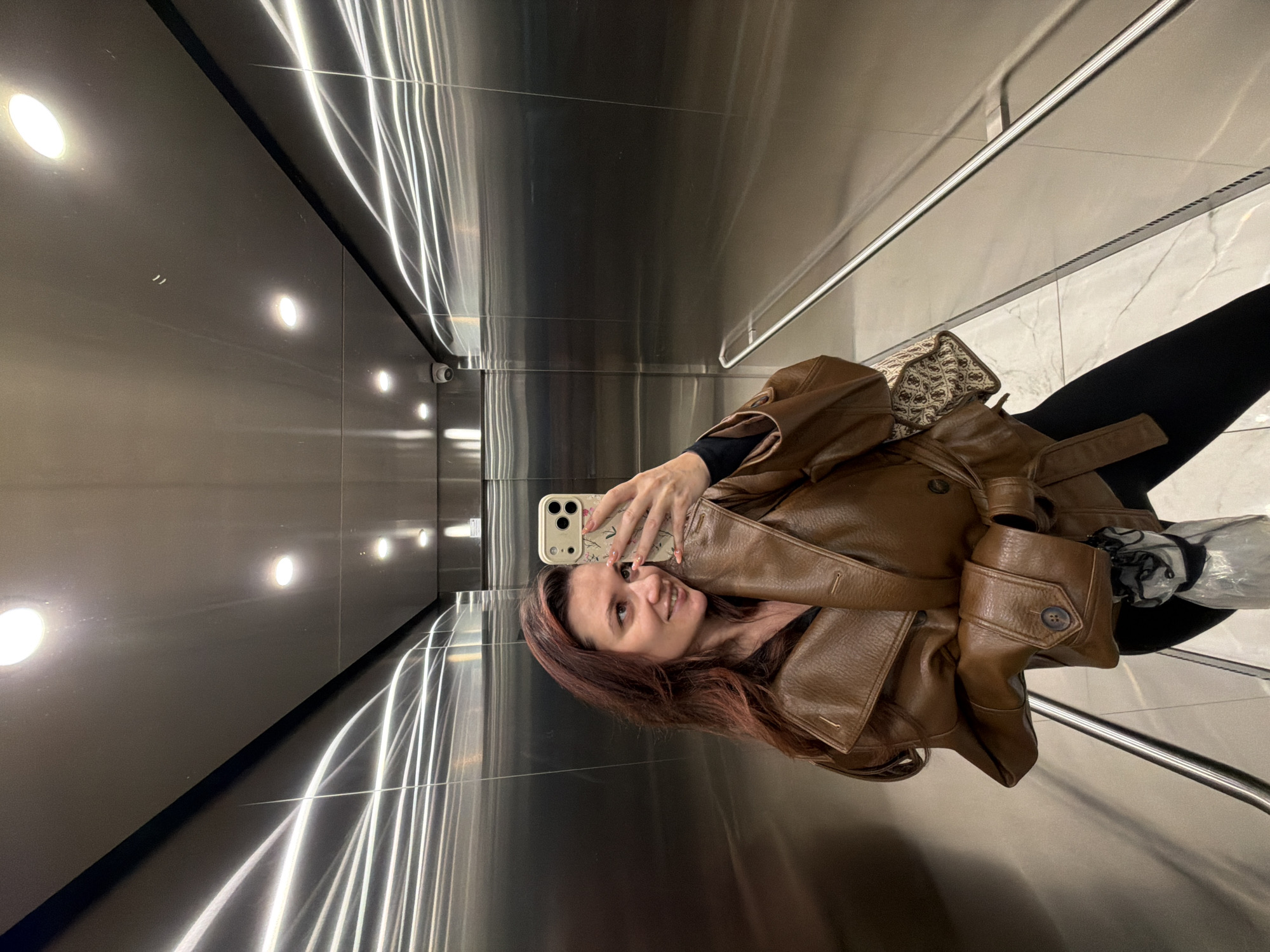 A woman is taking a selfie in an elevator. The walls of the elevator are stainless steel. There are recessed lights in the ceiling and shiny streaks reflecting off the stainless steel. The floor is tiled with white marble. The woman is wearing a brown leather jacket and black pants. Her hair is reddish-brown. She is holding a cell phone with a floral design case. The location cannot be determined from the given image.