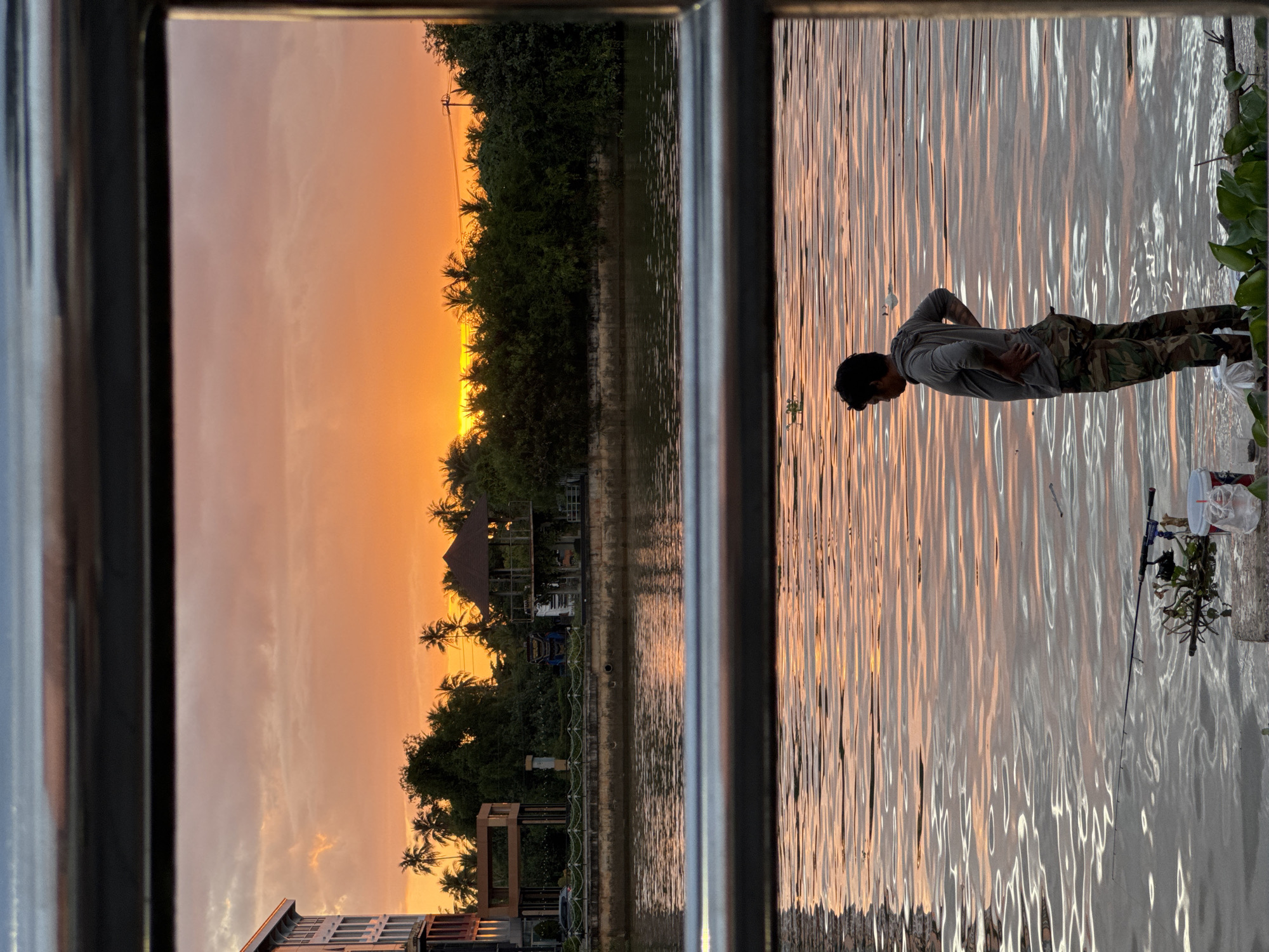 The image depicts a serene sunset scene at a waterfront in Surat Thani City Municipality, Thailand. In the foreground, a man stands on the bank of a river, facing away from the camera, with his hands on his hips. He is wearing a grey long-sleeved shirt and camouflage pants. A fishing rod is set up on the bank near him, with a white bucket and some greenery. The river's surface reflects the warm orange and pink hues of the sunset sky, creating gentle ripples.
In the background, across the river, is a waterfront promenade lined with lush green trees, palm trees, and some buildings. One building on the left appears to be a multi-story hotel. Further down the embankment, there is a small pavilion-like structure and a wooden frame. The sky is dominated by a vibrant sunset, with shades of orange, yellow, and pink clouds. Two horizontal metallic bars frame the top and middle sections of the image, suggesting the view is from a balcony or a similar vantage point. The overall atmosphere is peaceful and tranquil.