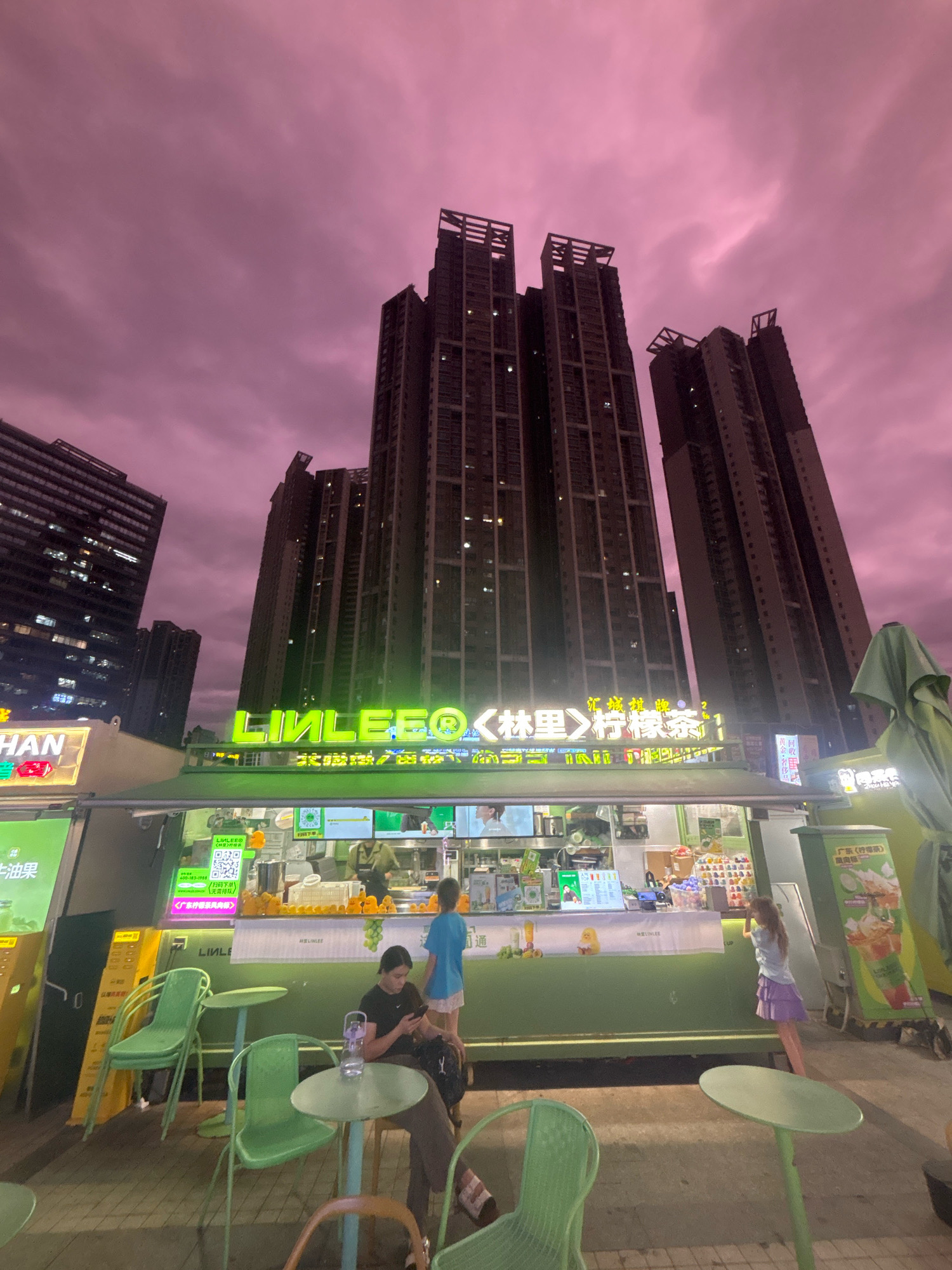 This is a low-angle, wide shot of an outdoor food stall in Nanhai District, China, with a backdrop of tall residential buildings under a pinkish, cloudy sky. The food stall is brightly lit and appears to be a beverage shop with green siding and neon green signage that reads "LIMLEE" and Chinese characters. Several people are visible. In the foreground, two individuals are seated at small green tables, one with a phone and the other looking away. A child in a blue shirt and purple skirt stands at the counter, possibly ordering. Another person, presumably an employee, is visible behind the counter, preparing drinks. The area around the stall has several empty green chairs and small tables. The overall scene suggests an evening setting, with artificial lights illuminating the stall and the surrounding area, contrasting with the dusky sky.