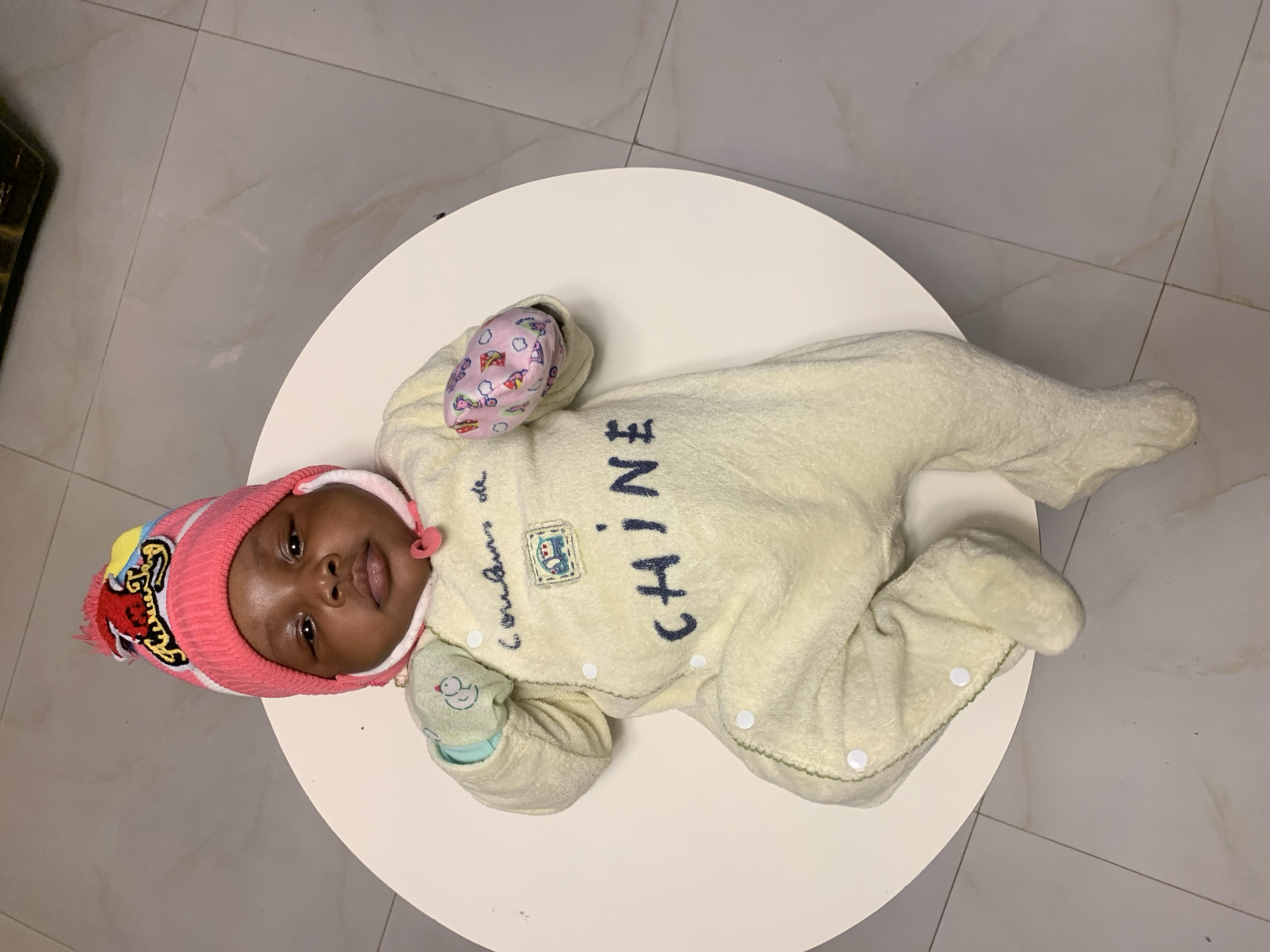 An infant is shown lying on its back on a round, white table, positioned in an indoor setting in Rafin Bauna, Nigeria. The baby has dark skin and is looking directly at the camera with a calm expression.
The baby is dressed in a light cream or pale yellow full-body onesie, possibly made of a soft, plush material. On the front of the onesie, the word "CHINE" is visible vertically in dark blue capital letters, and to its left, the cursive text "Couleurs de" is embroidered, accompanied by a small blue and white patch. The onesie features white snap buttons along the legs. The baby is wearing a pink knitted hat with a pom-pom and various colorful patches or patterns on the side. Pink mittens with a whimsical pattern are on one hand, while a light green mitten with a small white duck pattern is on the other.
The white table is placed on a tiled floor that appears to be light grey or beige with faint marbling patterns and dark grout lines. A dark, out-of-focus object is partially visible in the upper left corner of the frame. The lighting suggests a well-lit indoor environment, possibly daytime. The baby's attire, including the warm onesie, hat, and mittens, suggests a need for warmth or protection.