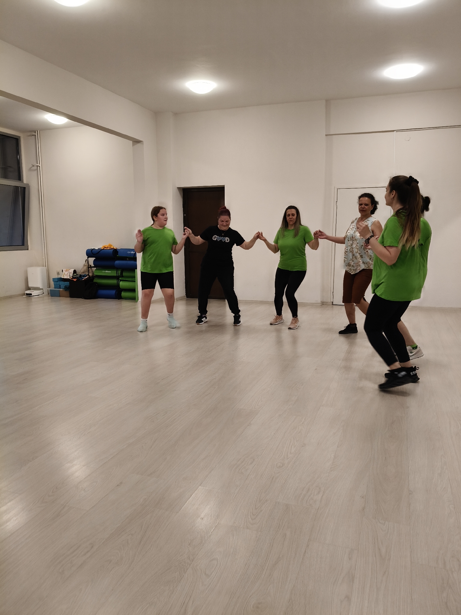 This full-shot image captures six women participating in a group activity within a brightly lit indoor studio in Plovdiv, Bulgaria. The women are holding hands, forming a loose arc or circle, and appear to be in motion, suggesting a dance or exercise class.
From left to right, the participants include a young woman in a green t-shirt and black shorts, followed by a woman in a black t-shirt bearing the text "GOOD" and black pants. Next is a woman in a green t-shirt and black leggings, holding hands with another woman wearing a light-colored patterned top and brown shorts. The group continues to the right with two more women, both in green t-shirts and black leggings; one's shoe shows the text "ESSE". Their expressions are generally focused and neutral, with some displaying slight smiles.
The studio features a light wooden floor and plain white walls. On the left side, windows are visible, and behind the initial participants, there are neat stacks of blue and green exercise mats or equipment. A white robotic vacuum cleaner is positioned near the stacked items. A dark brown door is visible in the background to the right of the equipment, with another door further right. The space is well-illuminated by multiple ceiling lights. The activity appears to be taking place in a dedicated fitness or dance area.