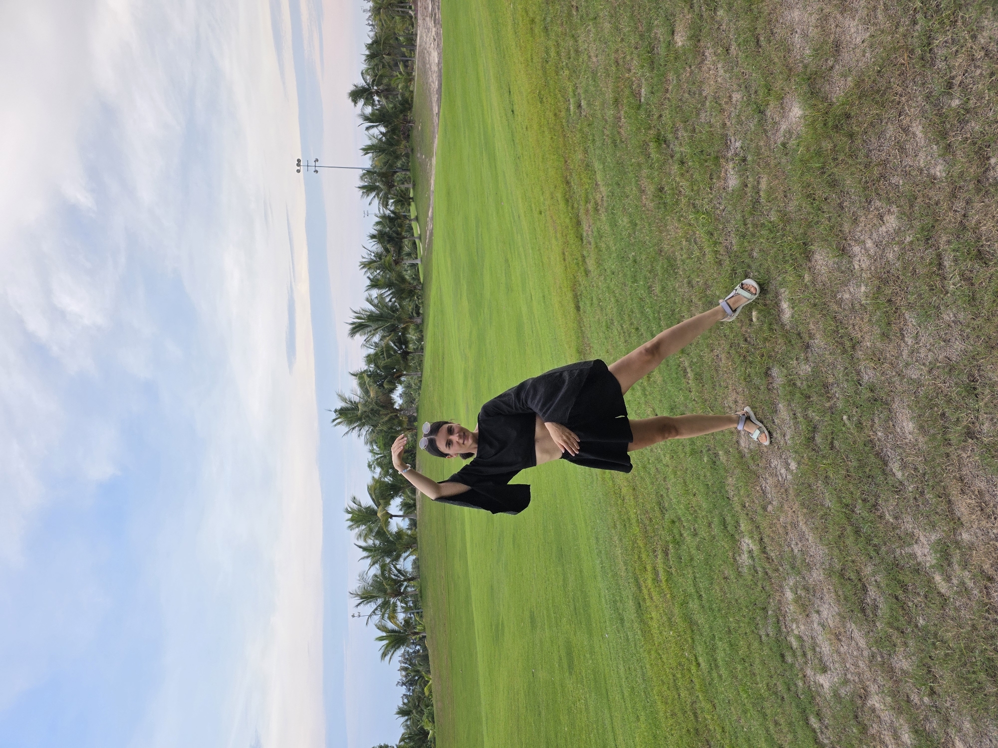 A full shot shows a female standing on a green and brown grassy area in Phu Thuy Ward, Vietnam. The female, fair-skinned with dark hair, is wearing a black crop top and black skirt, and white sandals. She raises her left arm and has sunglasses on her head. Behind the grassy area are palm trees. Beyond the palm trees are light blue skies with white clouds.