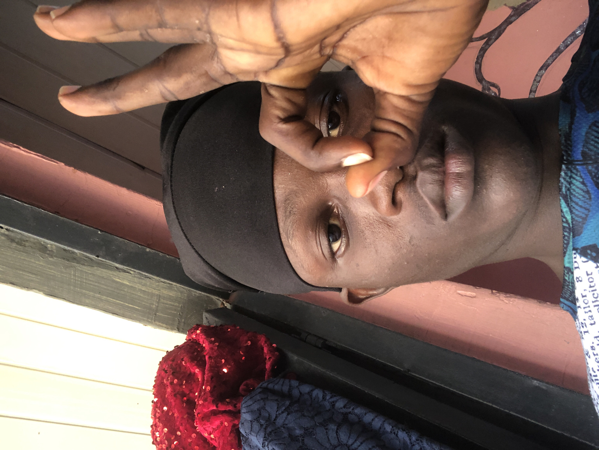 The image is a close-up selfie of a young Black male wearing a black headscarf. His right hand is raised, with his thumb and index finger forming a circle, obscuring his right eye. The background appears to be outdoors, with a pink wall, wooden slats, and some fabric items, including a red sequined cloth and a blue lace fabric, visible to the left. The overall lighting suggests daylight. The image does not contain any explicit information or visual cues to determine its location in Ado, Nigeria.