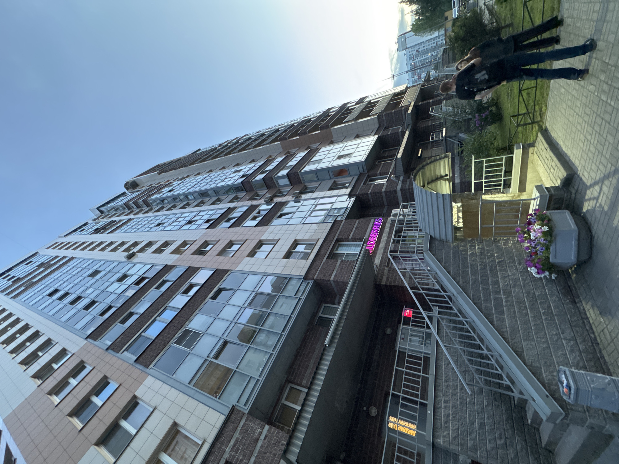 The image shows a modern, high-rise apartment building located in the city of Sinyavino, Russia. The building features a facade with a combination of beige panels and dark brickwork, interspersed with numerous large glass windows and balconies. A neon sign with pink lettering, partially visible, is mounted on the building's lower facade, above a set of stairs. Two individuals, a man and a woman, are standing on a paved walkway in front of the building, facing away from the camera. The man is wearing a black t-shirt and blue jeans, while the woman is wearing dark clothing. A gray stone planter filled with colorful flowers is positioned near the walkway. The sky above is clear and blue. In the background, other buildings and trees are visible.