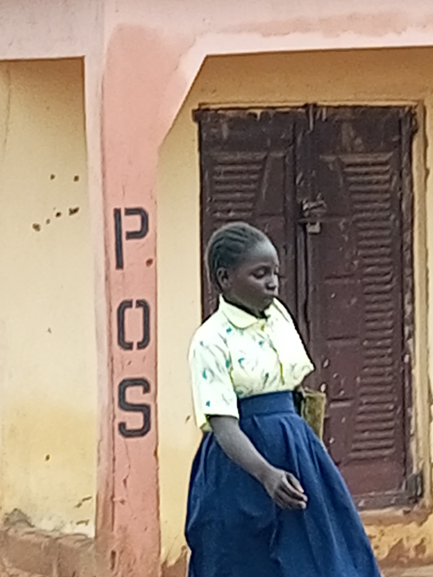 A young girl, appearing to be of African descent, stands in front of a building with a pink-painted pillar bearing the stencil markings "P.O.S." in black letters. The pillar is attached to a yellow wall. Behind the girl is a dark brown, louvered door. The girl is wearing a light-colored shirt with a patterned design and a dark blue, full skirt. Her left arm is bent at the elbow, and her hand is near her waist. She is looking down and to her left. The environment suggests an outdoor setting, likely in Gombe, Nigeria, given the style of architecture and the "P.O.S." marking which commonly denotes a Point of Sale, often used for financial transactions in that region.