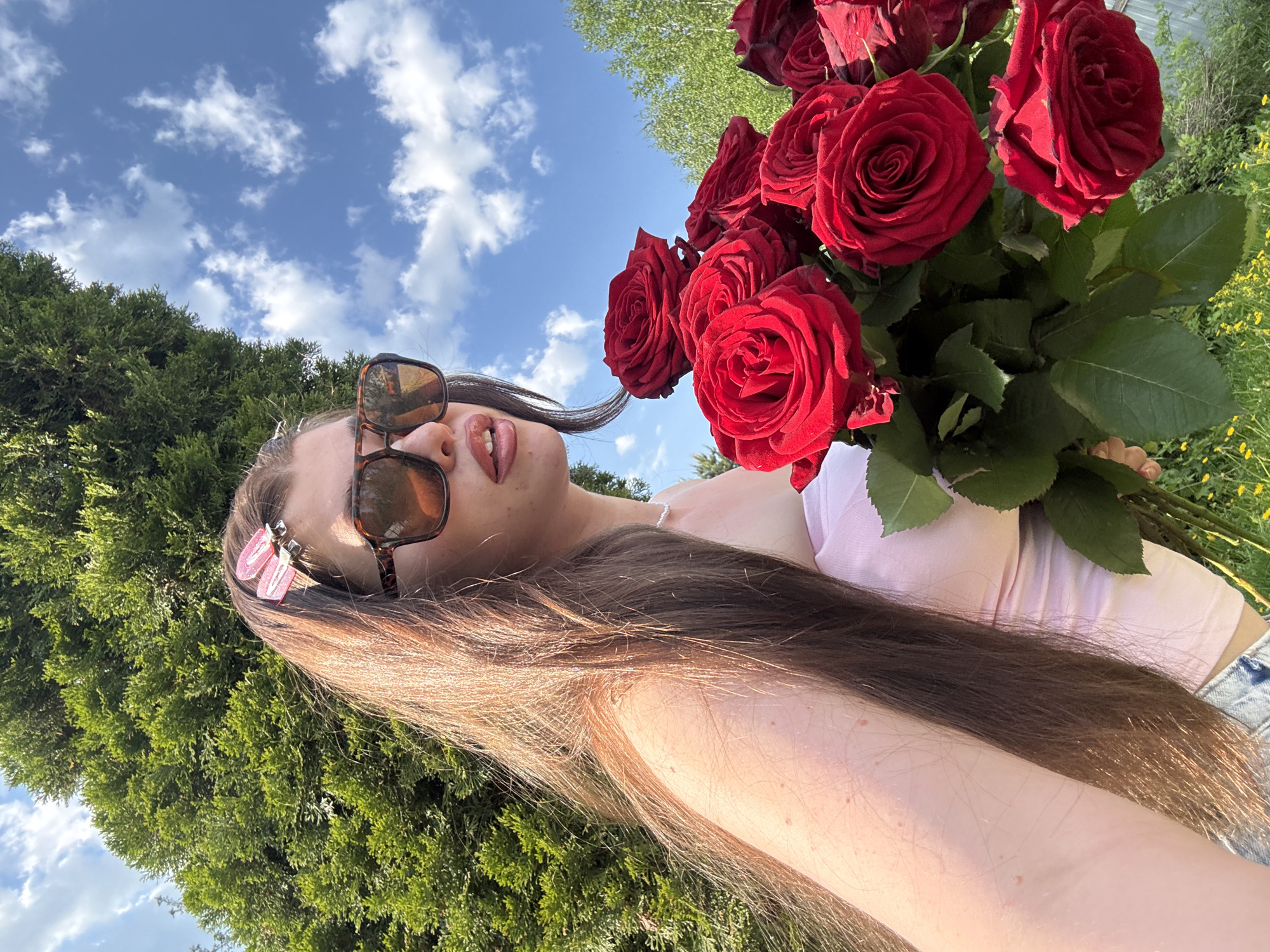 A young woman with long brown hair and sunglasses is holding a large bouquet of red roses outdoors. She is wearing a pink top and blue jeans. The background includes a blue sky with white clouds, green trees, and foliage. The photograph appears to be taken from a low angle. No information is available about the location in the city of Мещёры, Russia.