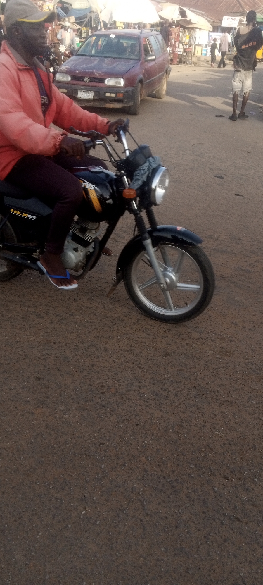 A man wearing a red jacket and a tan baseball cap is riding a black motorcycle on a street. The motorcycle is a TVS HLX 150. The man is wearing a pair of blue and white flip-flops. In the background, there is a maroon Volkswagen station wagon. Several other people are visible in the background, along with some market stalls. The image was taken in Narayi, Nigeria.