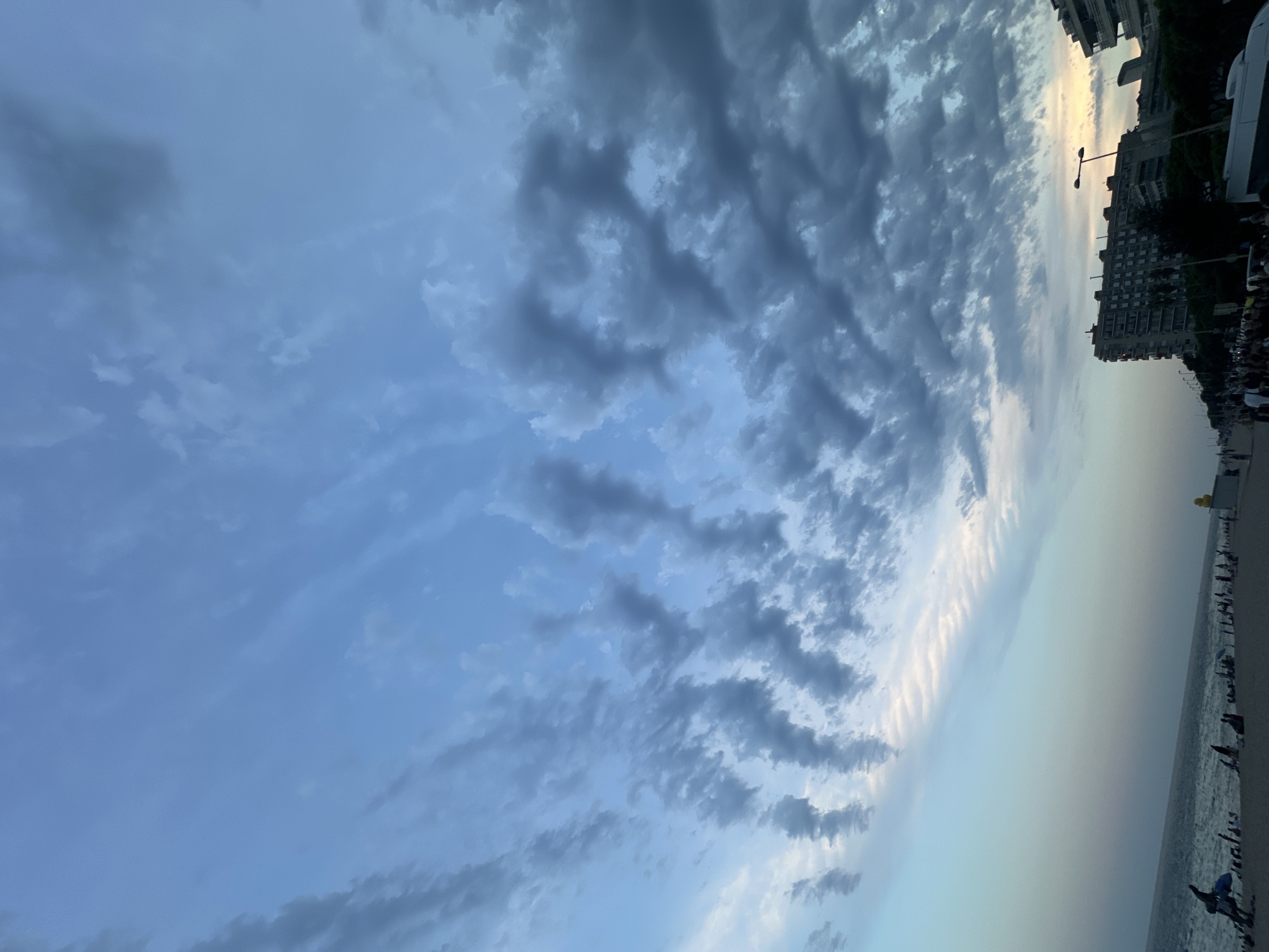 A vertical, slightly elevated shot captures a beach scene in Blanes, Spain. The sky dominates the upper portion of the frame, filled with dramatic, wispy grey clouds that contrast with patches of lighter blue and a faint, warm glow near the horizon. The lower portion of the image shows a wide beach lined with people, beach umbrellas, and some buildings in the distance. The sea is visible on the left, meeting the shore. The overall impression is of a late afternoon or early evening on a populated coastline.