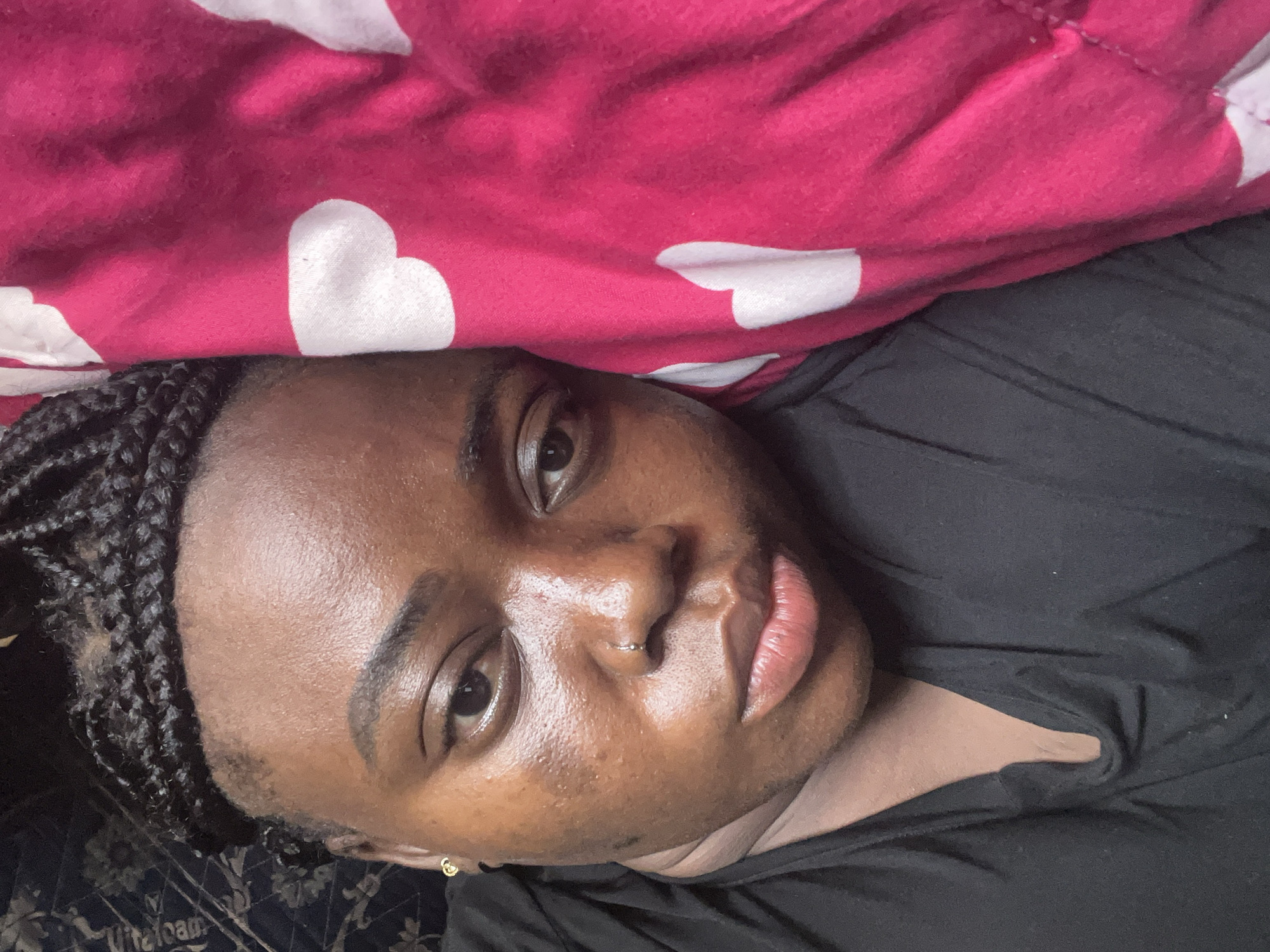 A close-up, slightly tilted portrait features a young Black person, likely female, lying down and gazing directly into the camera. The subject's head and upper torso are positioned diagonally across the frame. They have a dark complexion with a natural, slightly luminous finish, and maintain a calm, neutral expression. Their hair is styled in dark braids, visible on the left side of their head. A small, silver-colored septum ring adorns their nose, and a tiny gold-colored stud is visible in their right earlobe.
The person is dressed in a simple black garment, possibly a t-shirt or top, which covers their chest and right shoulder. Above their head, a vibrant pink blanket or comforter with large white heart patterns dominates the upper portion of the image. Below the subject's left side, a dark, subtly patterned fabric, possibly a sheet or pillowcase, is visible. Faint white text reading "Vitefoam" is discernible on this lower fabric.
The scene appears to be an indoor setting, likely a bedroom, indicated by the bedding. The overall impression is that of a personal, casual portrait or selfie. The brand name "Vitefoam" visible in the image serves as a location cue, as Vitefoam is a known mattress and foam manufacturer in Nigeria, consistent with the provided context of Abuja, Nigeria.