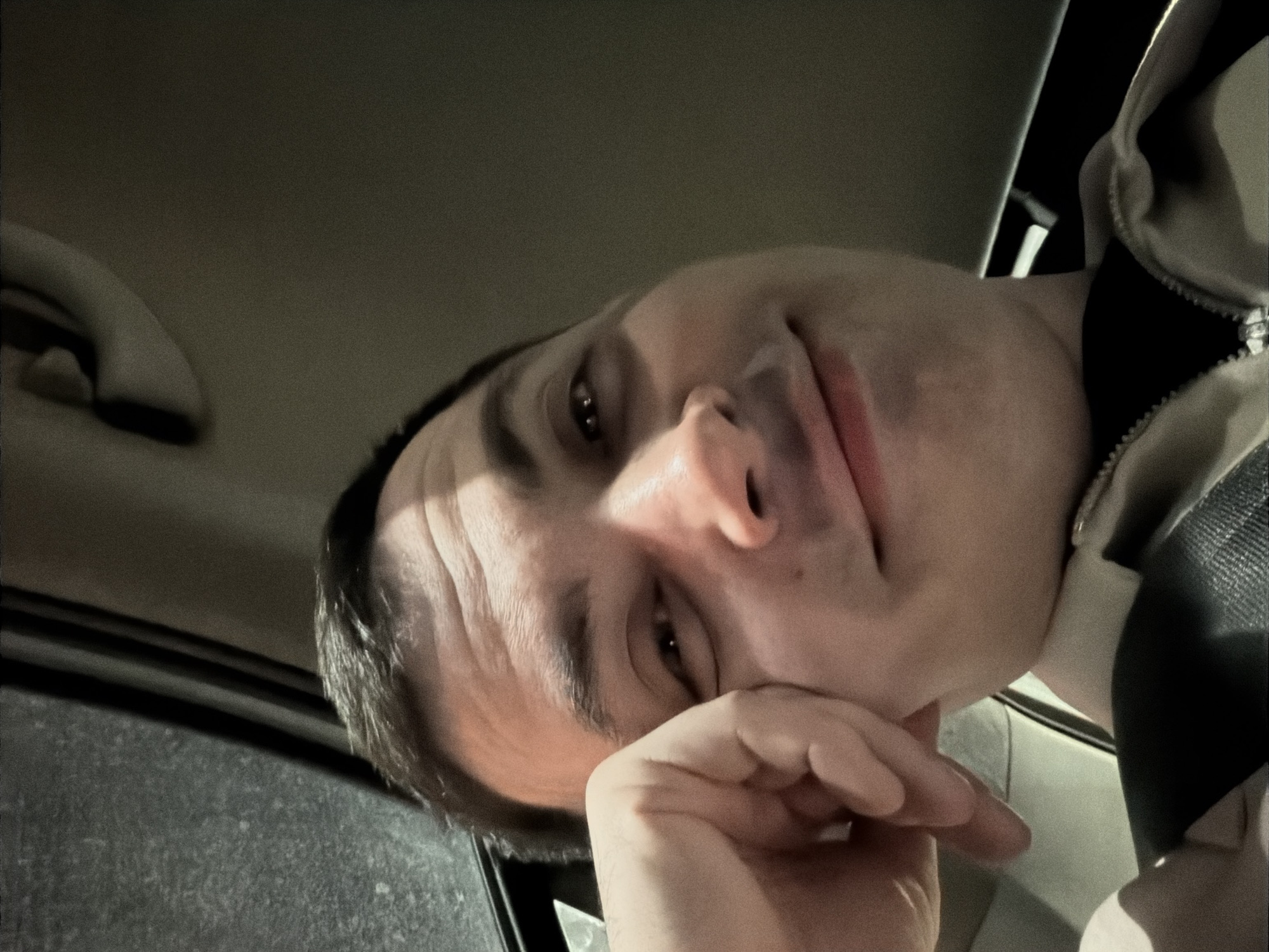 A person is in a car, possibly a sedan, with a light-colored interior. The person, a fair-skinned male, has short dark hair and is wearing a jacket with a visible zipper. The person's hand is resting on his face, partially obscuring it. A seatbelt is visible across the person's chest. The environment appears dark, possibly at night. The location in Izhevsk, Russia cannot be determined from this image.