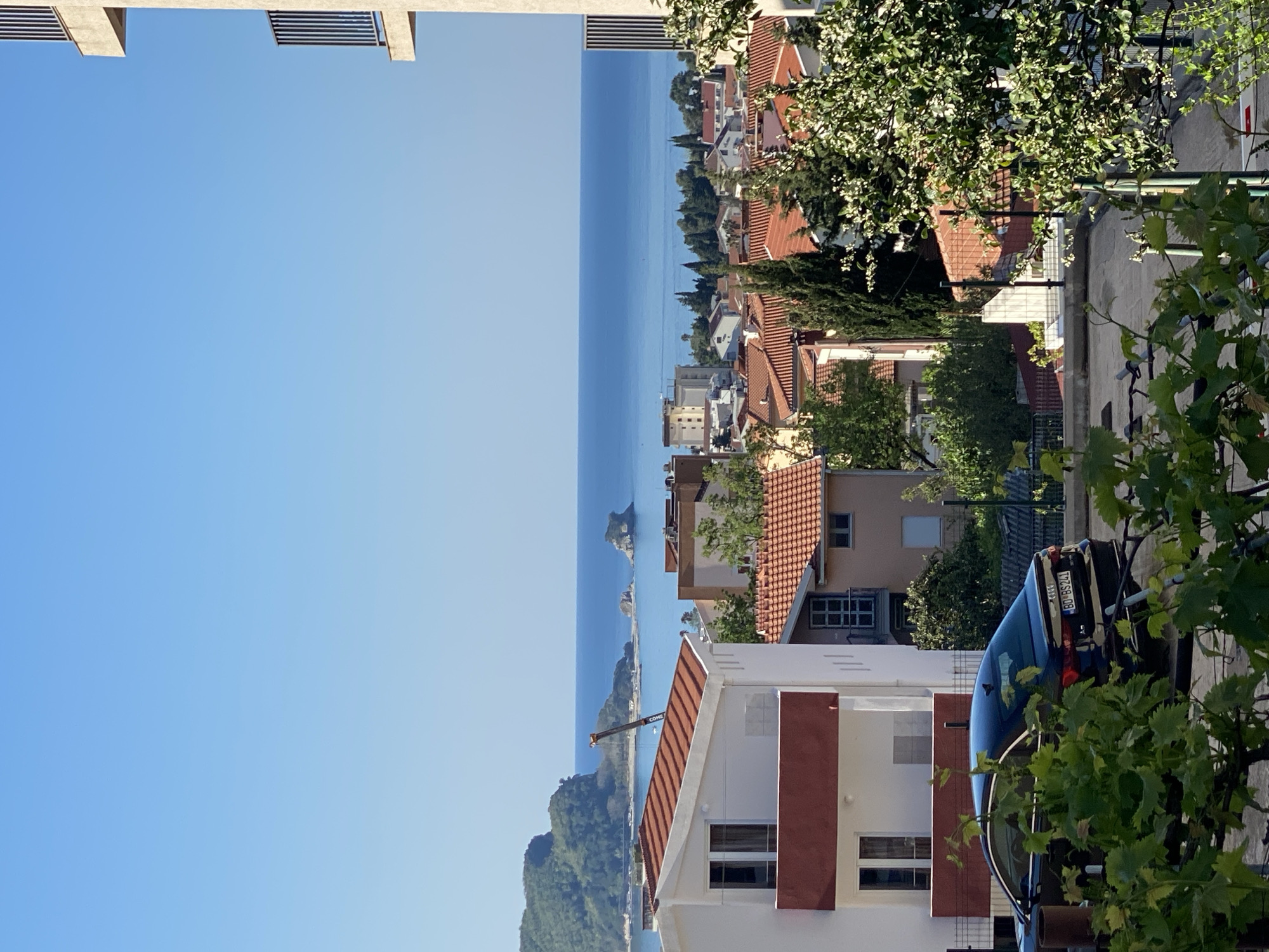 The image shows a view from an elevated position in Budva, Montenegro, looking out toward the Adriatic Sea. The foreground contains rooftops with terracotta tiles and a white building with brown accents. There are green trees and vine foliage framing the right side of the shot. In the middle ground, there are various buildings and structures situated along the coastline, creating a layered, urban-seascape composition. The sea stretches to the horizon under a clear blue sky. In the right corner is a car parked on the street.