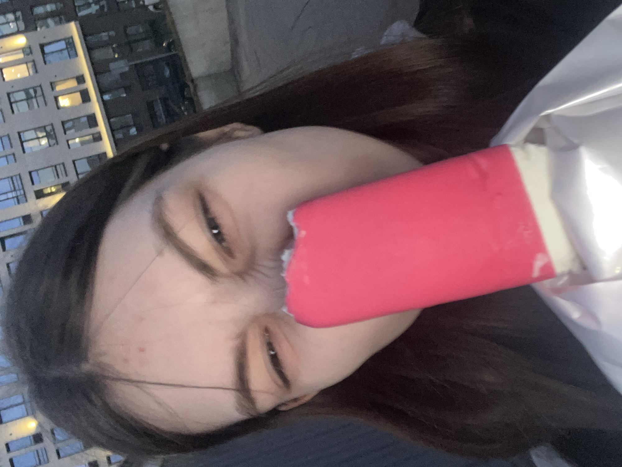 A young woman is eating a red popsicle, in front of a modern apartment building in the city of Saint Petersburg, Russia. The building has many windows and is made of gray concrete. The woman has brown hair and is wearing a dark shirt. The popsicle is partially eaten, with a bite taken out of it. It is wrapped in white paper.