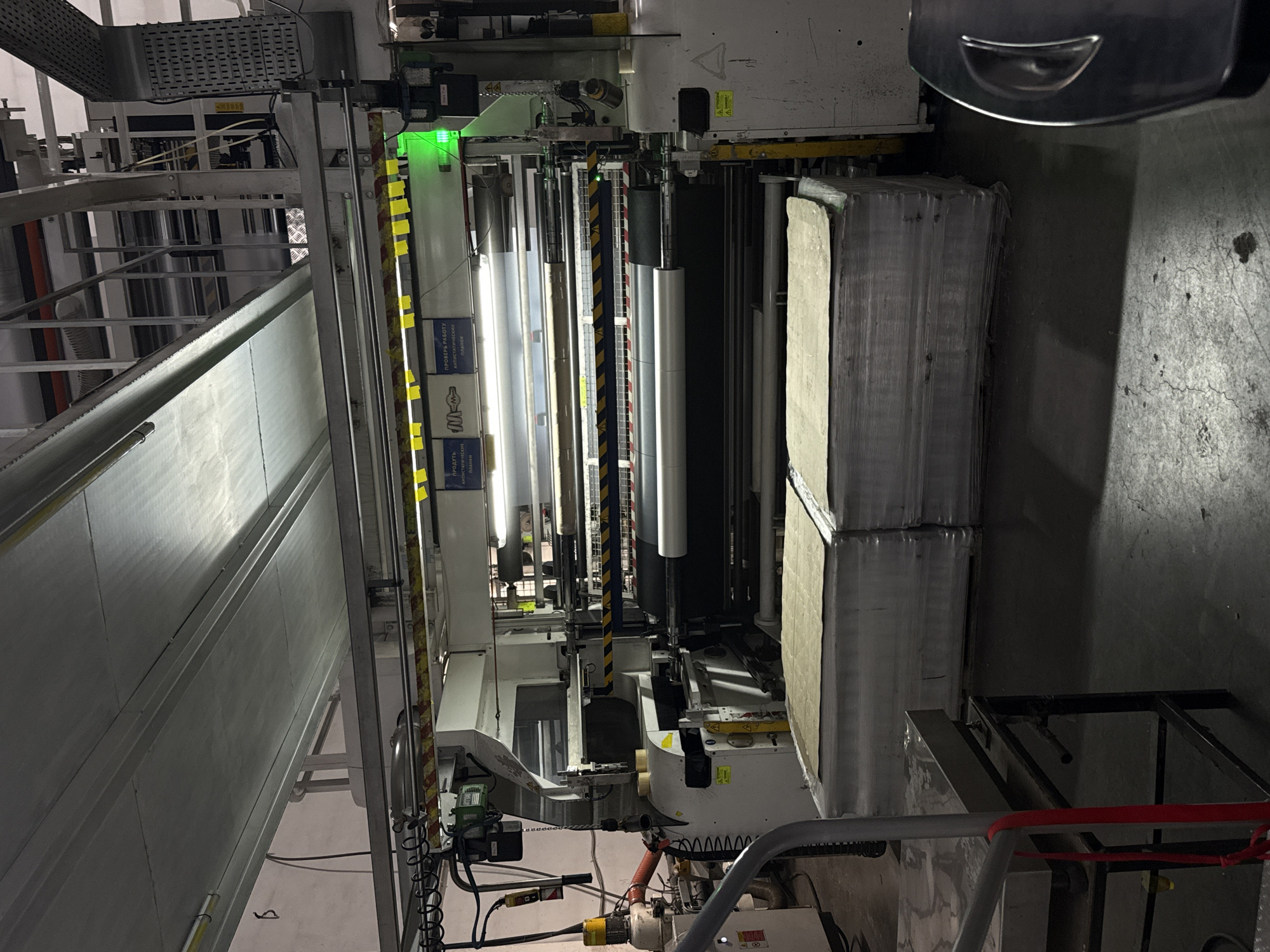 This image captures an interior view of an industrial manufacturing facility in Gamovo, Russia. The scene is dominated by a large, complex machine featuring multiple rollers, metal frames, and various components indicative of a production line, possibly for printing, converting, or processing sheet materials.
In the center, large, metallic rollers are prominent, with bright artificial lighting illuminating the area. A distinct green indicator light is visible on the upper part of the machinery, suggesting operational status or a system alert. To the right, stacks of what appear to be processed sheets or raw materials, possibly paperboard or similar goods, are neatly arranged and wrapped in plastic. The floor is concrete with visible cracks and stains, typical of an industrial environment. Overhead, an intricate network of metal structures, conduits, and wires forms part of the facility's infrastructure.
Visible text on some panels or boxes in the upper right part of the machine is in Russian. One reads "ПРОВЕРЬ РАБОТУ АНТИСТАТИЧЕСКИХ ПЛАНОК" (Check the operation of anti-static bars) and another "ПРОДАТЬ АНТИСТАТИЧЕСКИЕ ПЛАНКИ" (Sell anti-static bars). A logo that appears to be "М-М" is also present. Yellow and black striped warning tape is wrapped around some structural elements. The facility is under artificial lighting, with no natural light sources visible, suggesting it could be any time of day or night. No people are present in the frame, but the machinery implies ongoing or readiness for industrial activity.