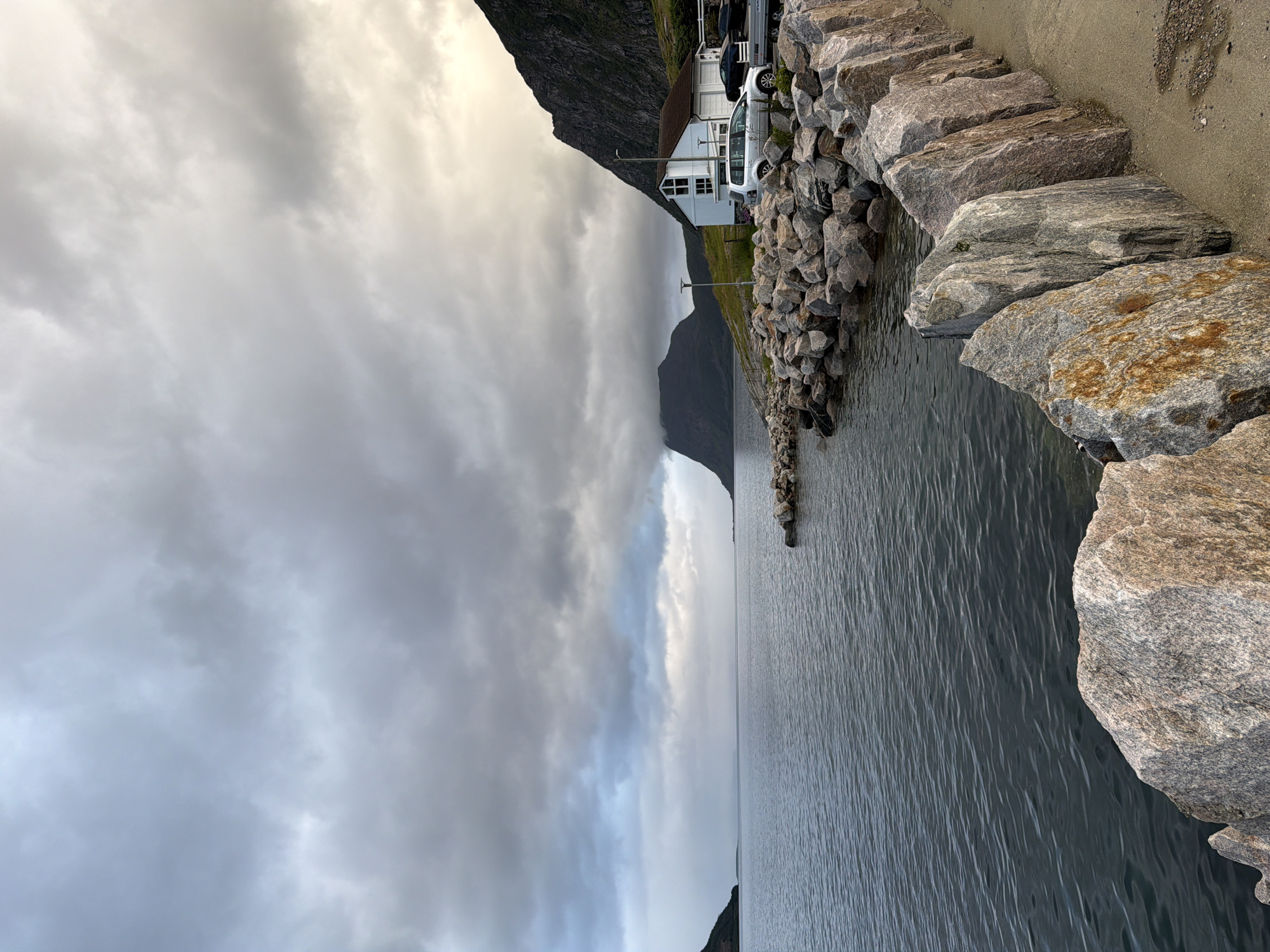 The image depicts a coastal scene with a body of water in the foreground and a dramatic mountain landscape in the background. The sky is overcast with thick, gray clouds, with a slight hint of warmer light breaking through on the right side.
On the right side of the frame, a white building with a brown roof is situated on the shoreline. In front of the building, several vehicles are parked, including a silver SUV and a dark sedan. The building appears to be a residential or possibly a small commercial structure.
The shoreline is fortified with a wall of large, irregular rocks, which extend from the right foreground into the water. These rocks are various shades of gray and tan. The water is calm, with gentle ripples visible.
Further in the distance, on the left side of the image, a distinctive, cone-shaped mountain rises from the water, partially shrouded in clouds. Another, larger mountain range dominates the right side of the background, with steep, rocky slopes. The overall impression is one of a rugged, northern coastal environment.
Based on the distinctive mountain shape and the architectural style of the building, this location is likely to be near Harstad, Norway, possibly along the coastline where such geological features and structures are common.