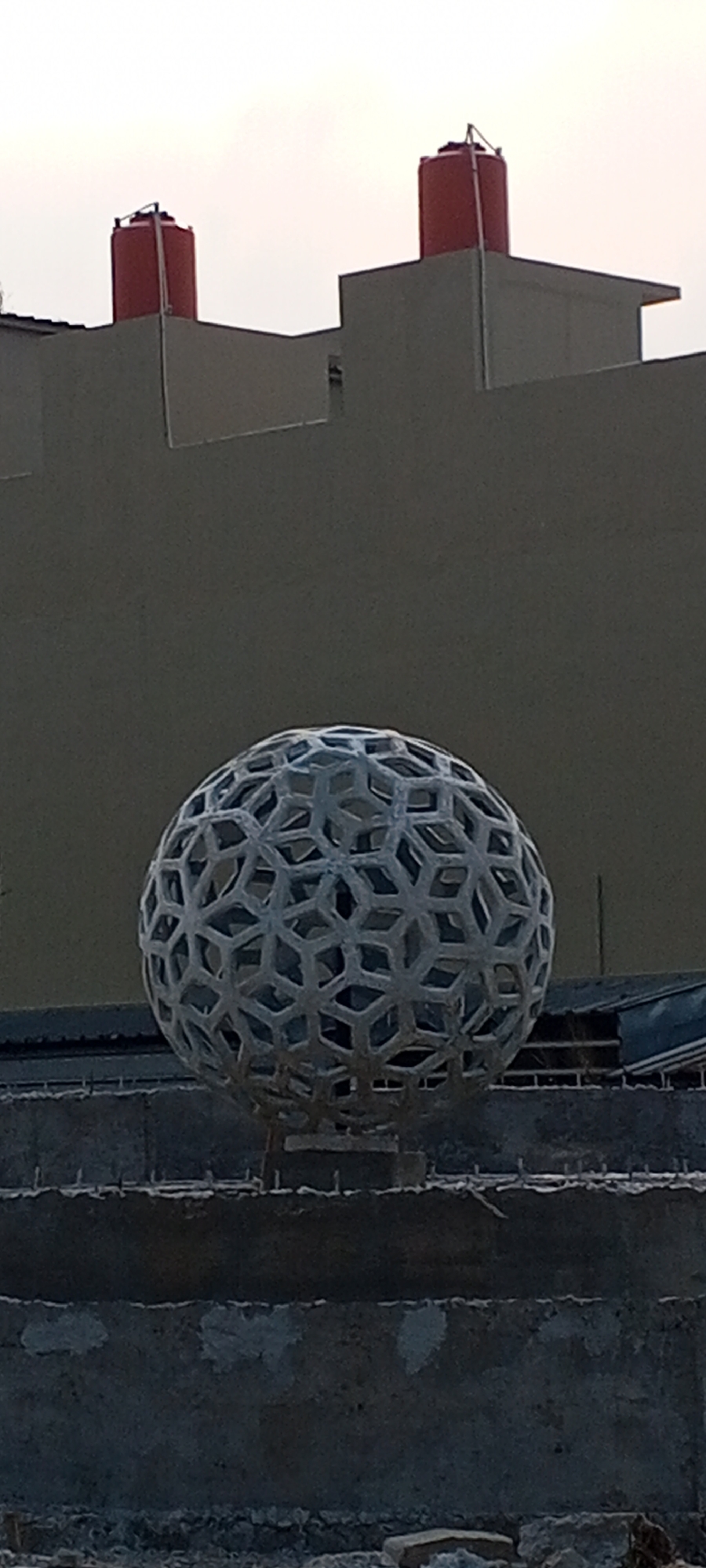 The image depicts an outdoor scene at what appears to be a construction site in Depok, Indonesia. In the foreground, a grey, spherical sculpture with a geometric, honeycomb-like pattern is centrally positioned. It is elevated on a pedestal of concrete blocks. Behind the sculpture, a dark grey building facade rises, topped with two cylindrical red water tanks. The sky above is a pale, overcast grey. The overall impression is one of urban development or renovation.