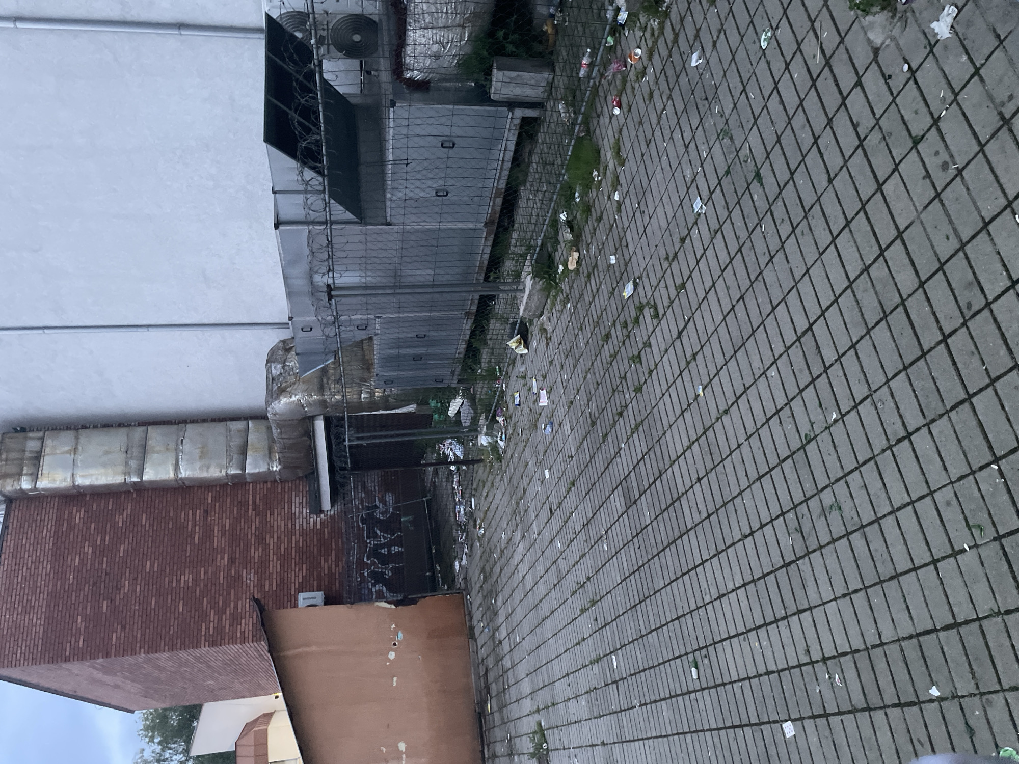 The image displays an outdoor, neglected area, likely a service or back alleyway, in Ihtiman, Bulgaria. The ground in the foreground and mid-ground is paved with grey rectangular concrete blocks, with patches of grass growing between them. This paved surface is extensively littered with various debris, including numerous plastic bottles, aluminum cans, paper scraps, plastic wrappers, and other indistinguishable refuse.
A tall metal fence, resembling a chain-link fence, runs horizontally across the mid-ground, acting as a barrier. The top of this fence is fitted with barbed wire or razor wire. Behind the fence, on the left side of the frame, are large industrial elements: a white building wall and metallic structures that appear to be air conditioning units or large ventilation ducts, one featuring a circular fan. To the right, behind the fence, a building with reddish-brown brick walls is visible, along with sections of a light brown-colored wall and more metallic piping or ductwork.
No people or activities are taking place; the scene is static. The lighting suggests an overcast day, possibly late afternoon or early morning, contributing to the dull overall atmosphere. The pervasive litter indicates a lack of maintenance or improper waste disposal.
On the brick wall, dark graffiti is visible, composed of stylized letters that are difficult to read, possibly "2XYX." Below this, a small white box-like object has faint, barely legible text that might spell "SHARPO" or "SHARP."