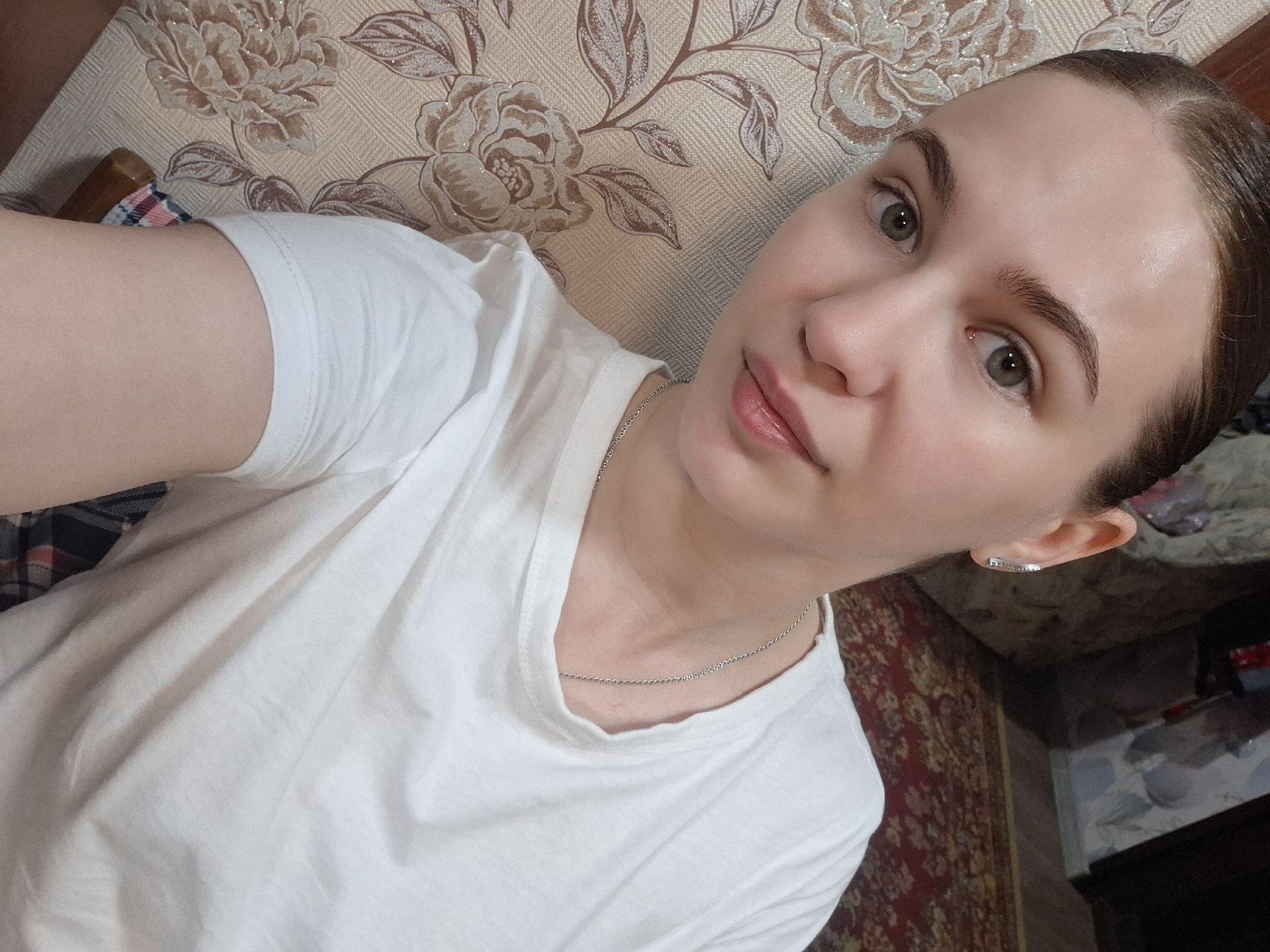 A person with fair skin and light brown hair pulled back is pictured in a selfie. The person is wearing a white t-shirt and a silver necklace. The background includes a floral-patterned wall and a red patterned rug. The location is Nizhnyaya Gar', Russia.
