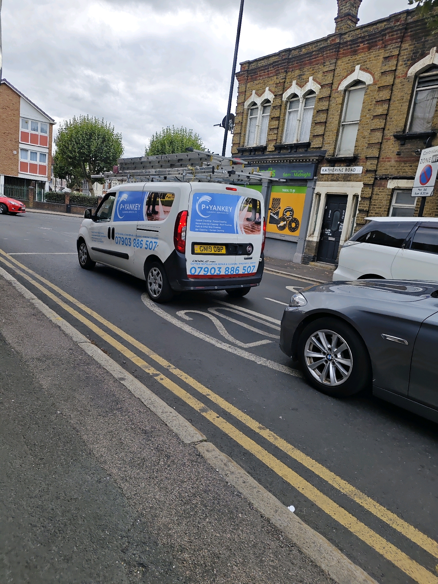 In the city of Greater London, United Kingdom, a white van with "P+Yankey Cleaning Services Ltd" and the phone number "07903 886 507" printed on its side is traveling on a wet road. The van has a ladder rack on its roof, indicating a cleaning or maintenance service. A grey BMW is in the foreground, partially obscuring the road. The background features a row of yellow brick buildings with arched windows and a sign that reads "Katherine Road". A red car is visible on the left side of the image. The sky is overcast, suggesting a cloudy day. The road has double yellow lines, indicating parking restrictions.